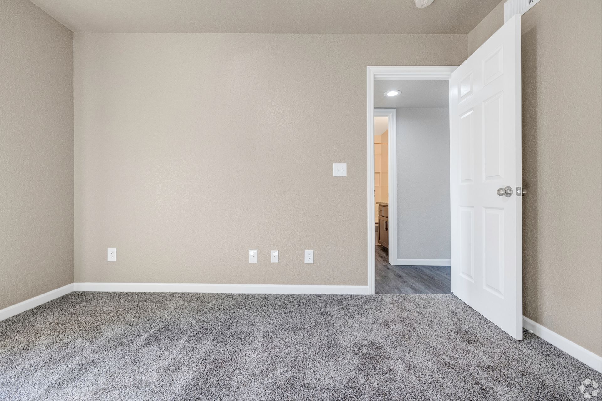 Empty room with gray carpet, beige walls, and an open white door leading to a hallway.