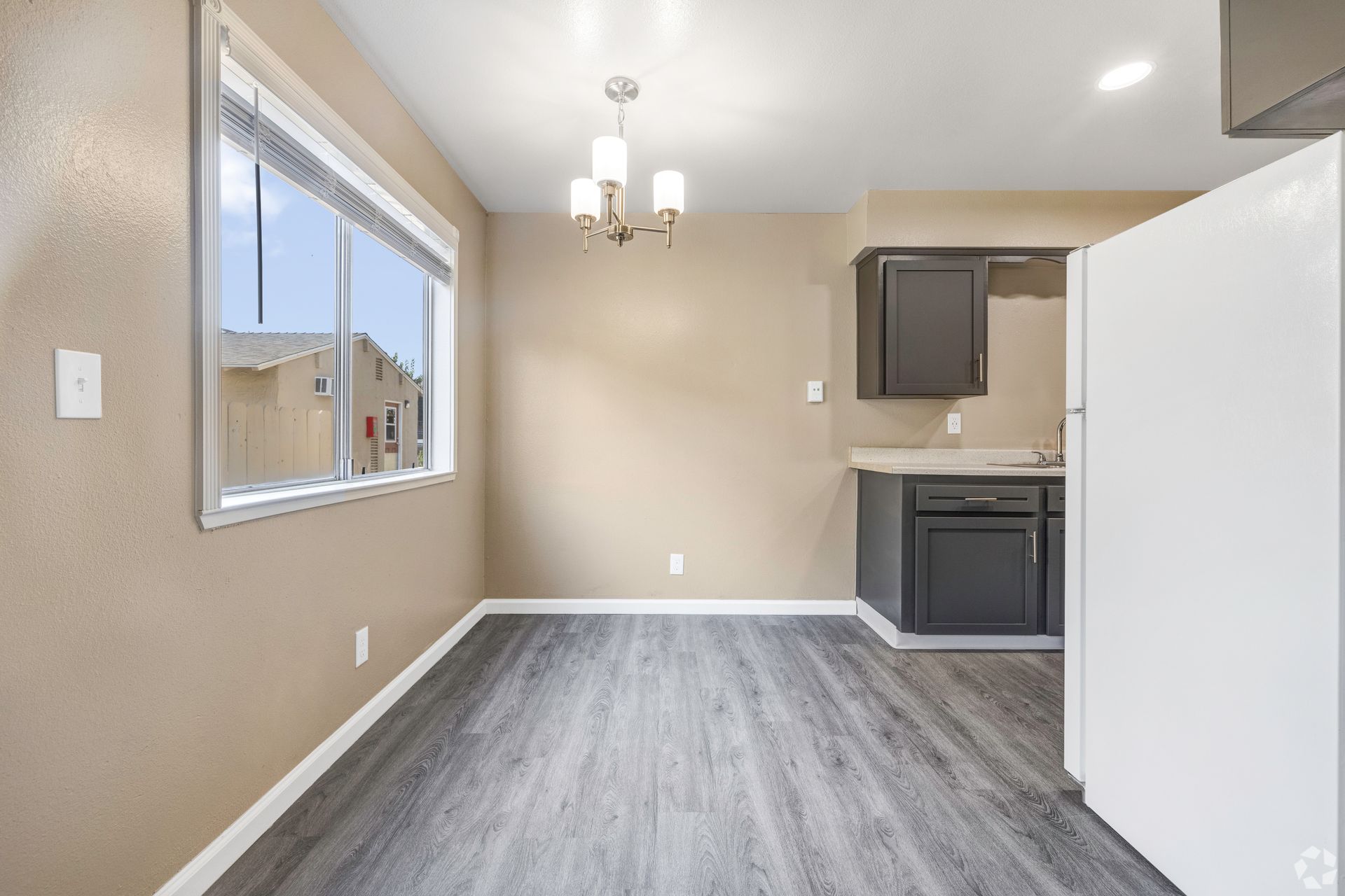 Dining area with gray floor and tan walls, window, chandelier, and small kitchen with dark cabinets.