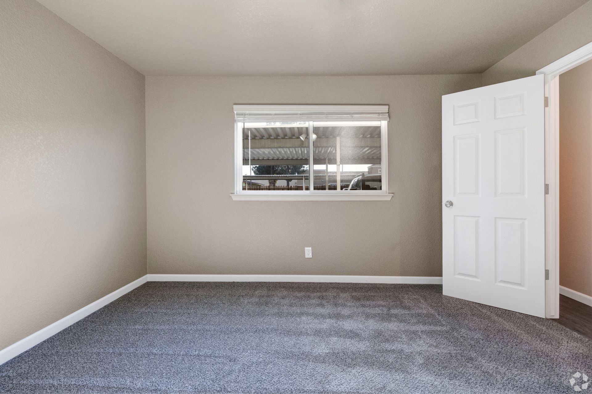 Empty bedroom with gray carpet, beige walls, window, and partially open white door.