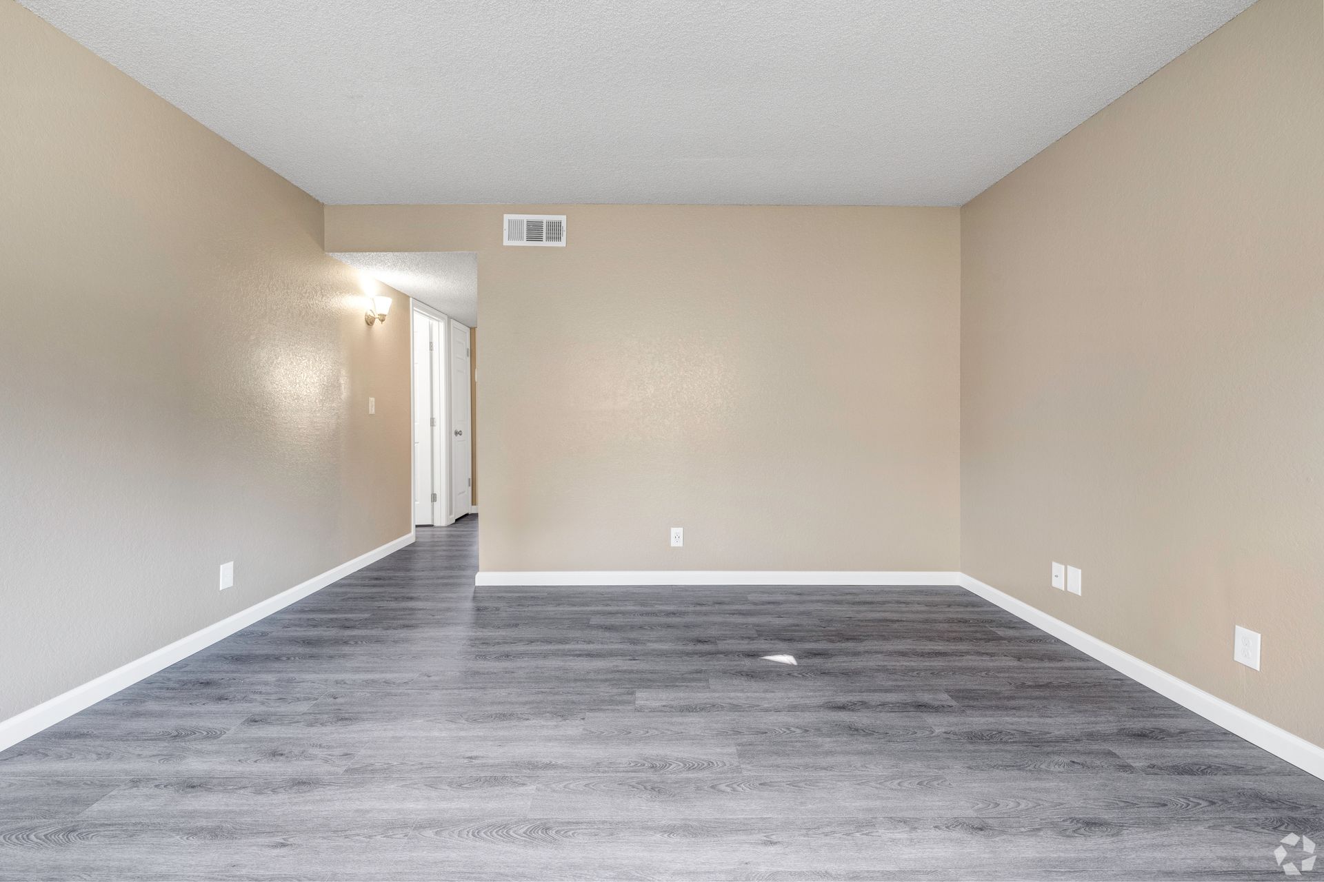 Empty living room with gray wood-look floors, beige walls, white trim, and a doorway to another room.