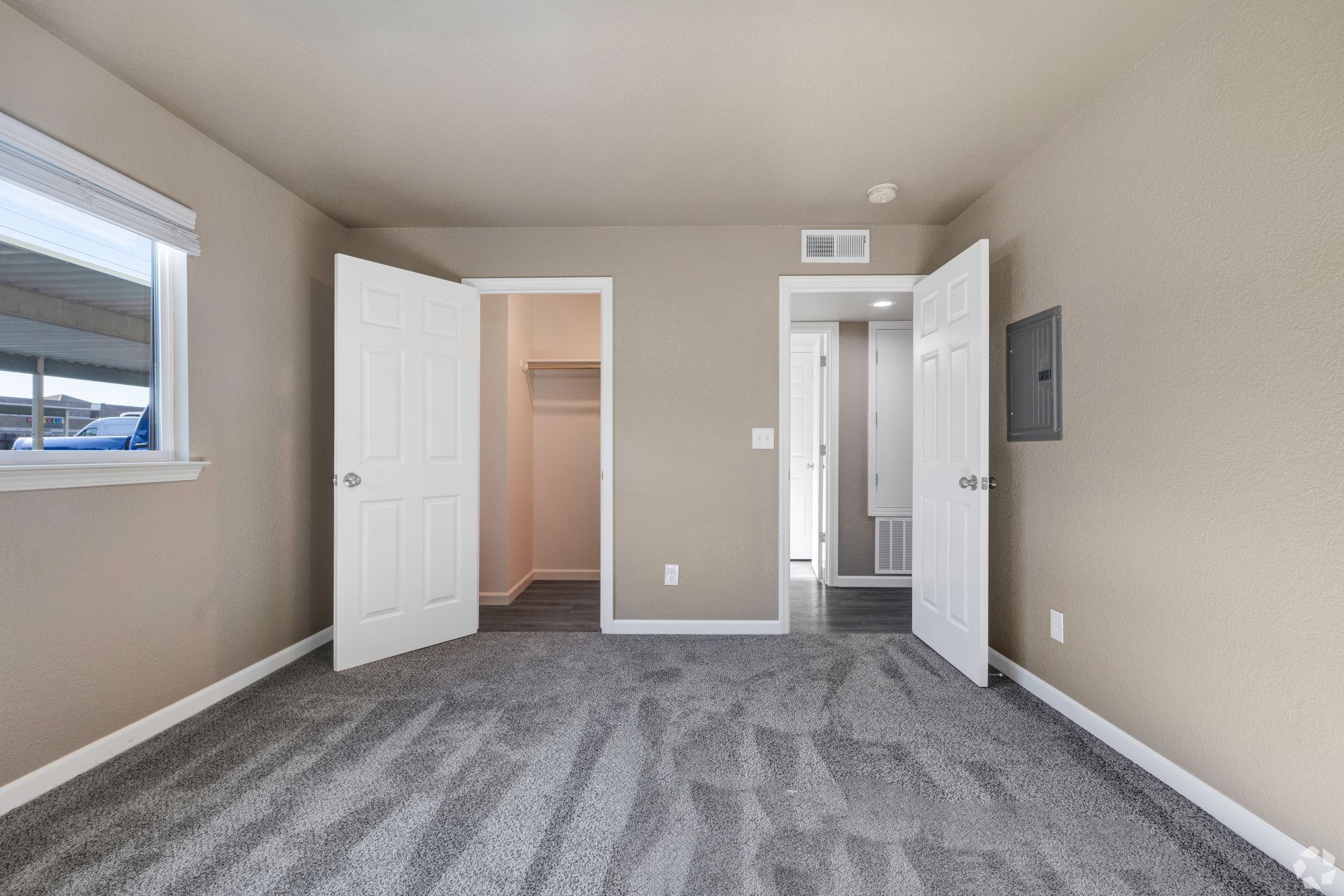 Empty bedroom with gray carpet, beige walls, two open white doors, and a window.