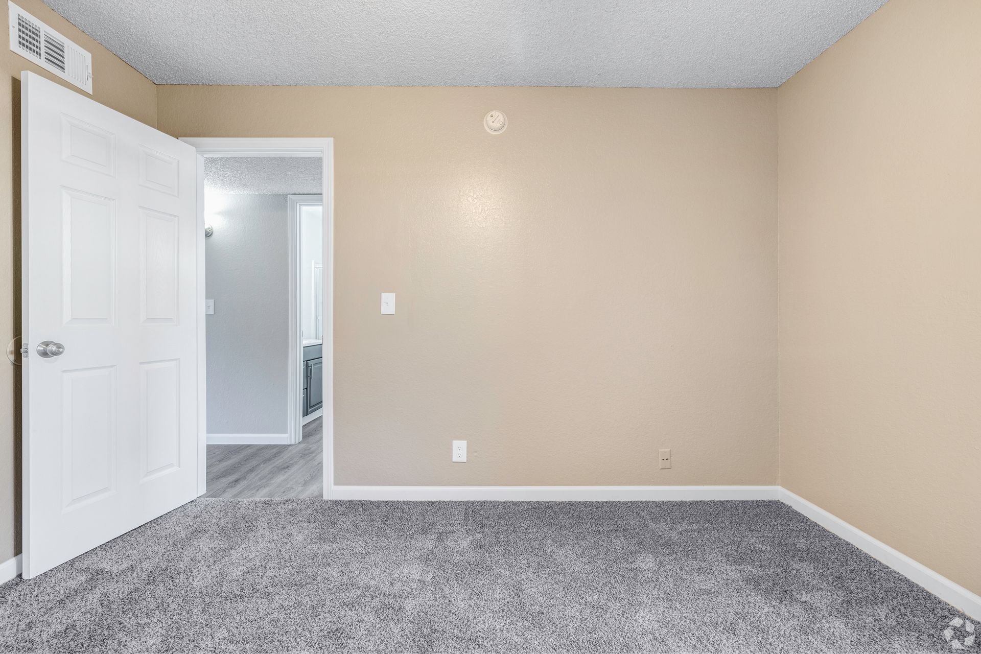 Empty room with beige walls, gray carpet, and an open doorway to another room.