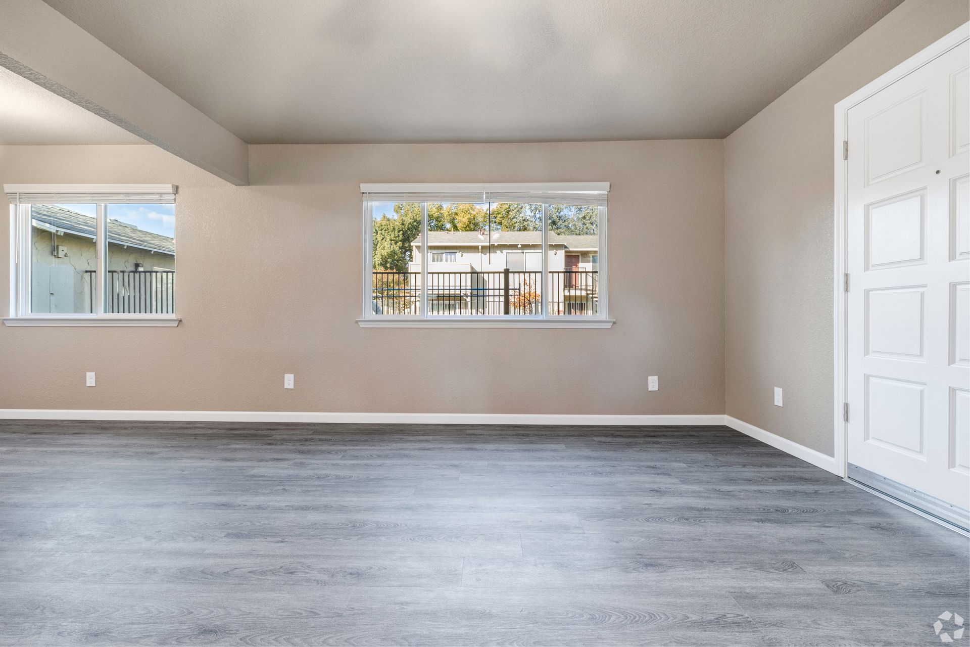 Empty living room with gray floor, beige walls, white door, and windows overlooking a neighborhood.