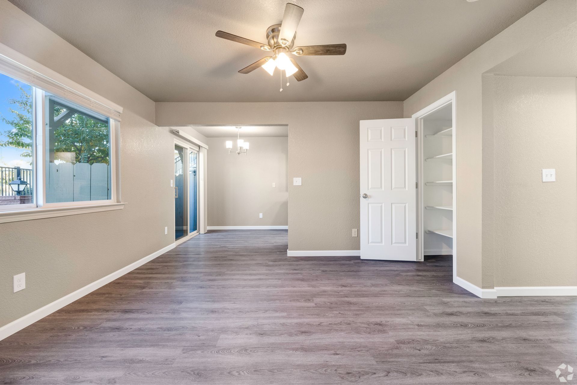 Empty living room with wood-look flooring, tan walls, a white door, and a ceiling fan.