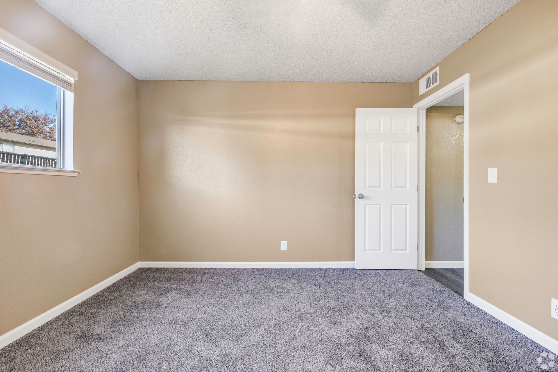 Empty bedroom with gray carpet, tan walls, white door and window.