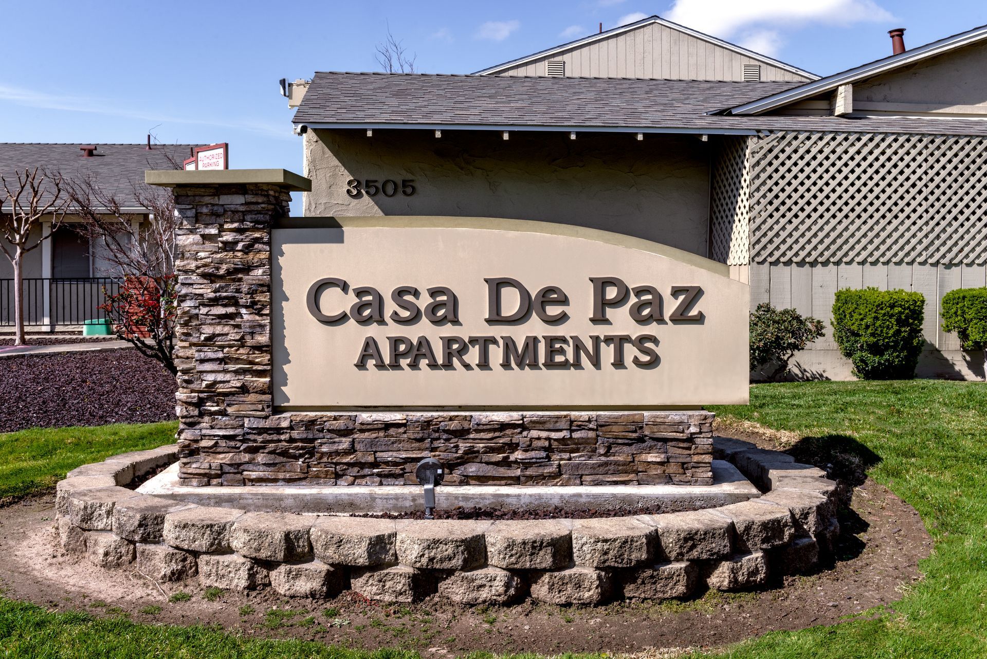 Casa De Paz Apartments sign in front of building, brown and tan with stonework, on a sunny day.