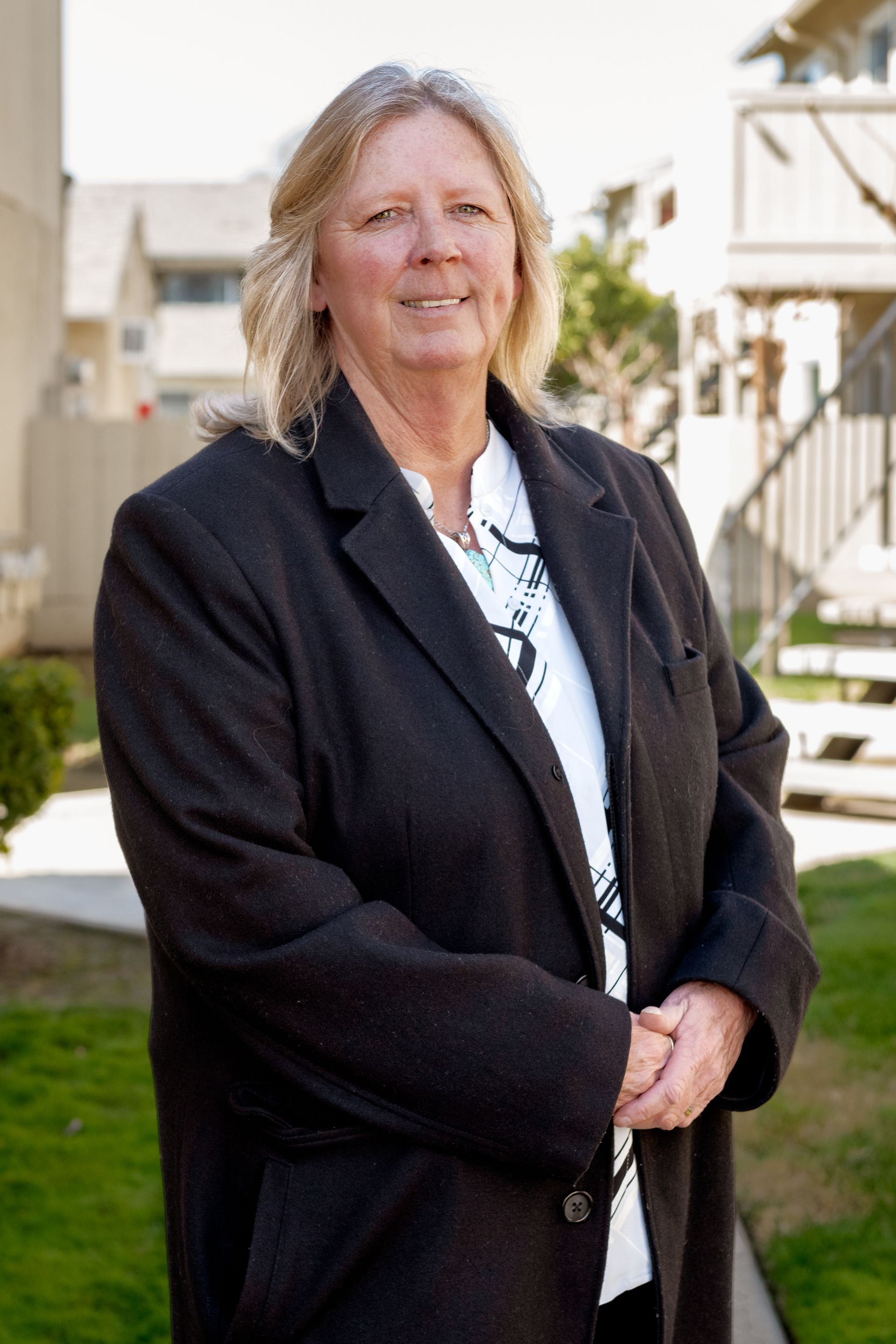 Woman with blonde hair, in a black coat and white blouse, standing outside, smiling slightly.