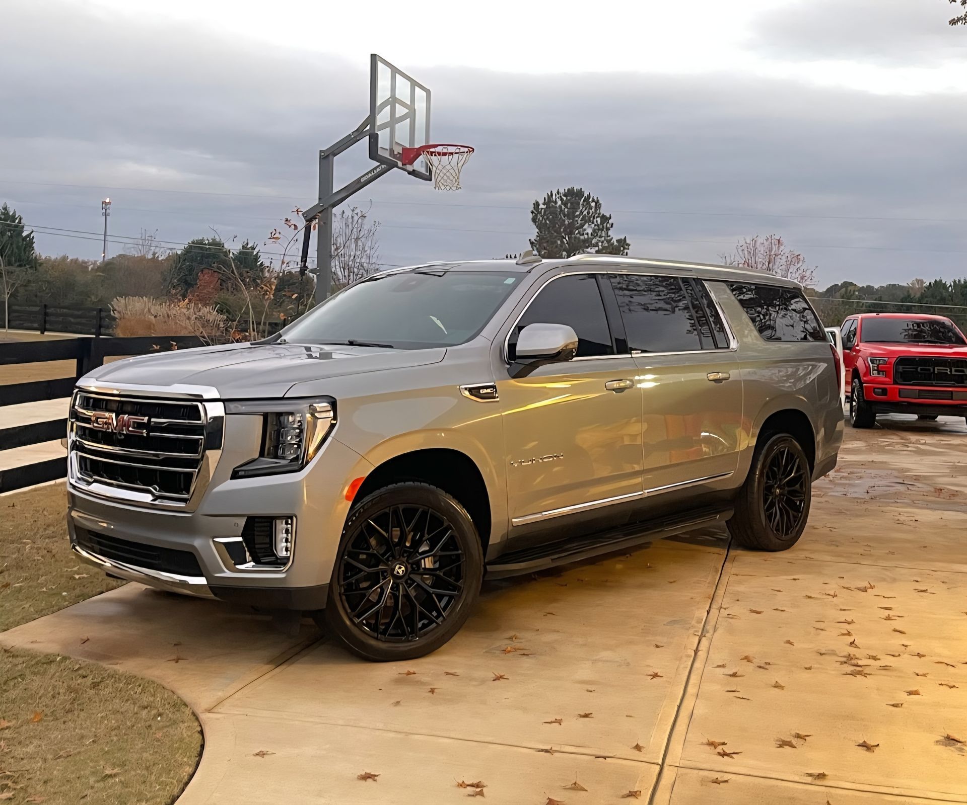 Silver SUV parked outdoors on a driveway beside a basketball hoop, with a red truck in the background.