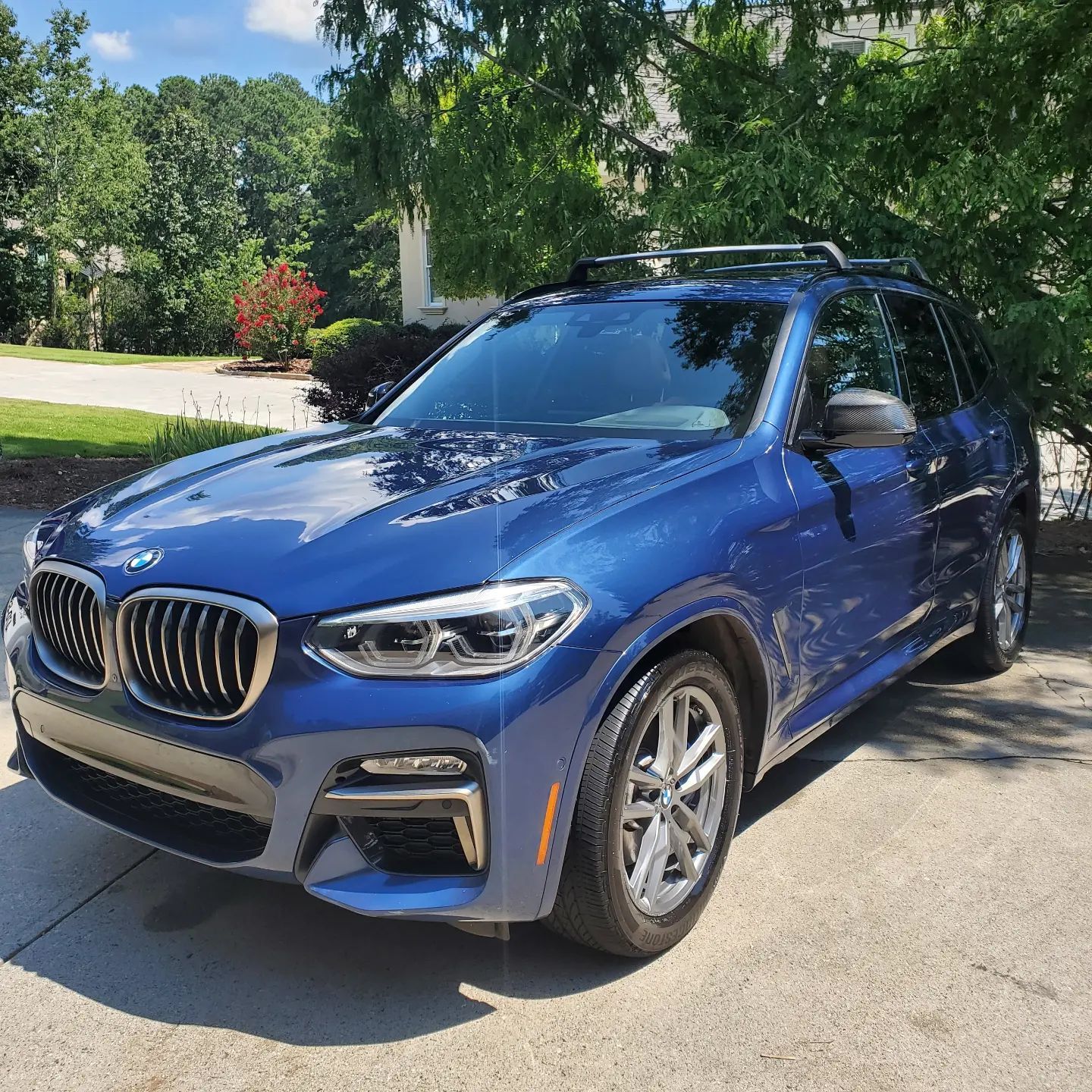 Blue BMW SUV parked on a driveway with a roof rack. Green trees and a house in the background.