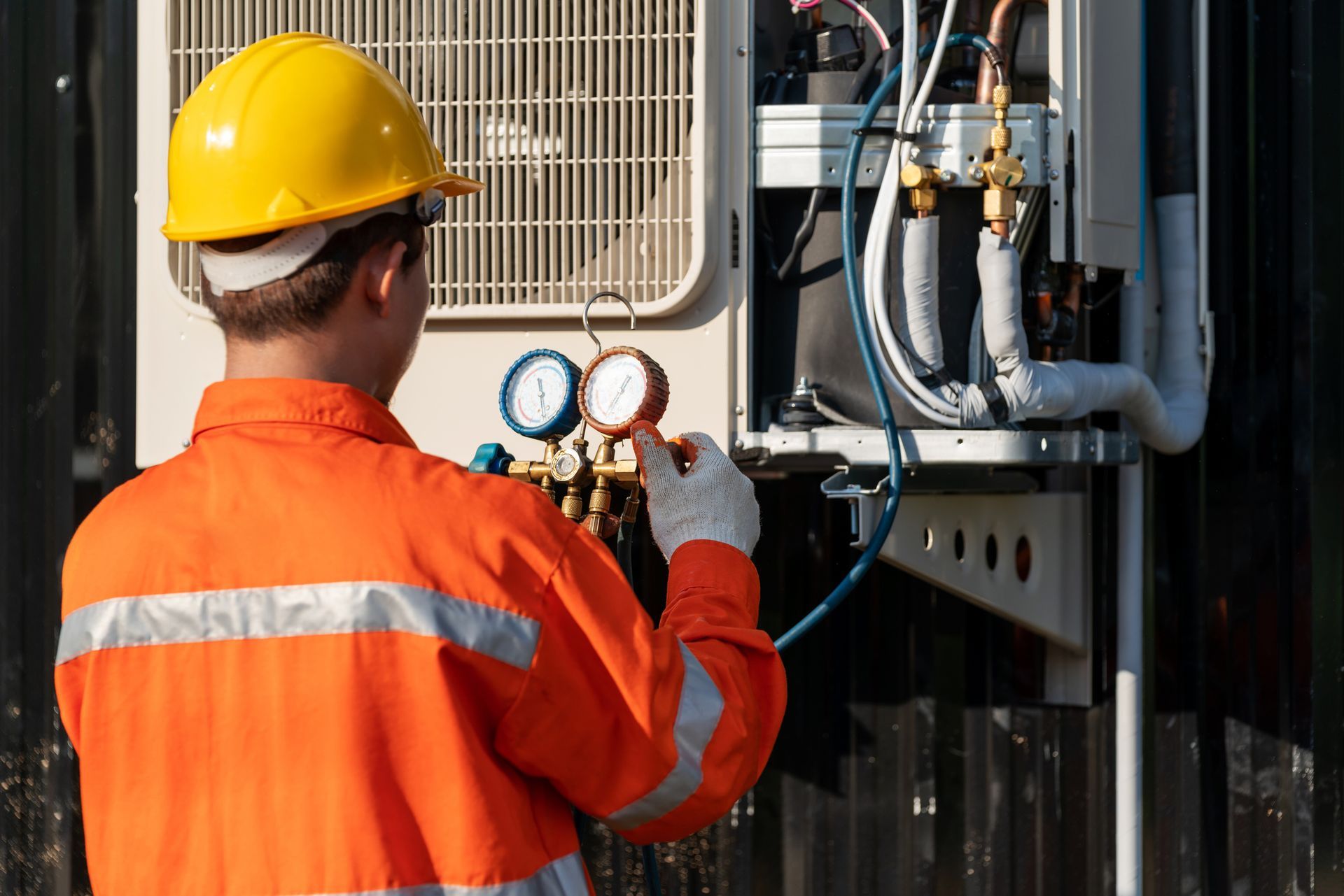 HVAC technician in orange uniform, yellow hard hat, checking gauges on an outdoor air conditioner unit.