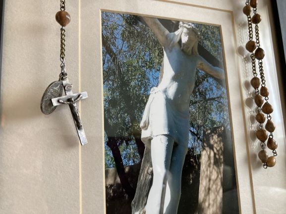 White statue in a framed outdoor scene, flanked by hanging rosary beads and a crucifix.