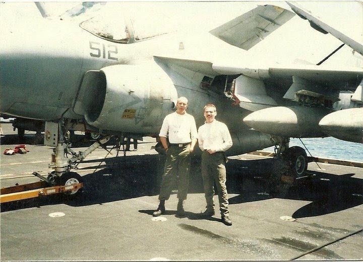 Two men stand beside a military jet on a ship deck, with aircraft in the background.