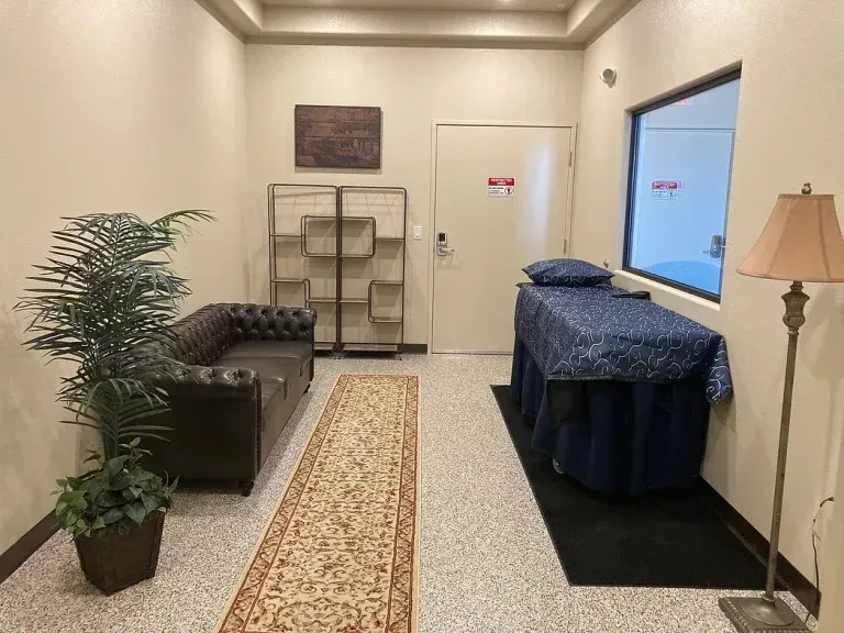 Small waiting room with black couch, shelving, massage table, floor lamp, and window.