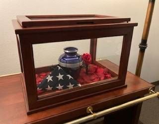 Glass display case with a blue vase and red flower atop a folded American flag on a wooden table