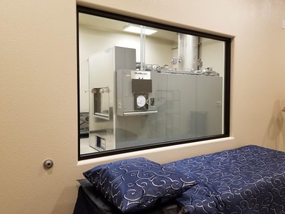 Hospital bed seen through a window into a lab or clinical room with equipment and cabinets.