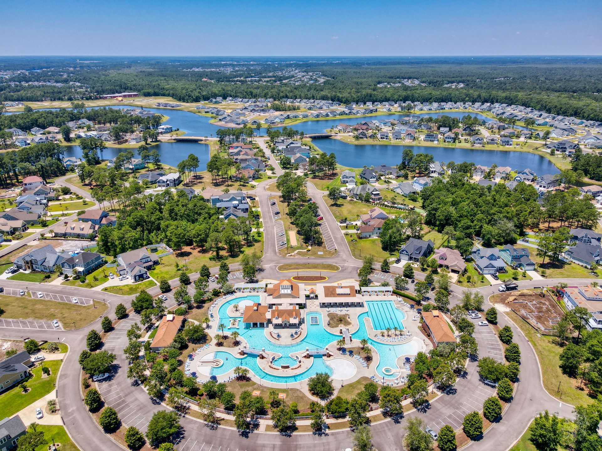 Aerial view of a community with a large pool complex, housing, and lakes. Green trees and blue water.