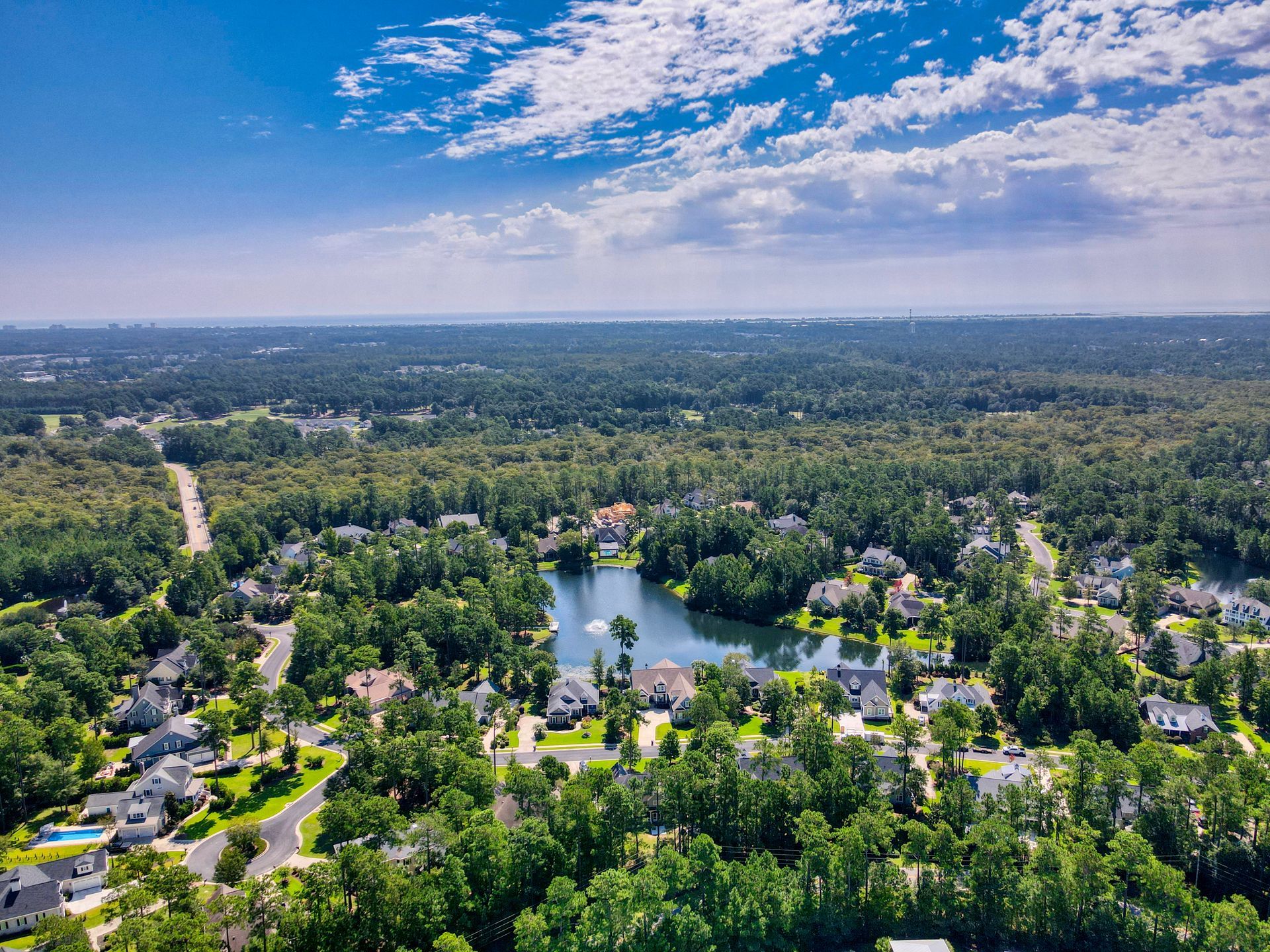 Aerial view of a suburban neighborhood with a lake surrounded by lush green trees and houses, under a blue sky.