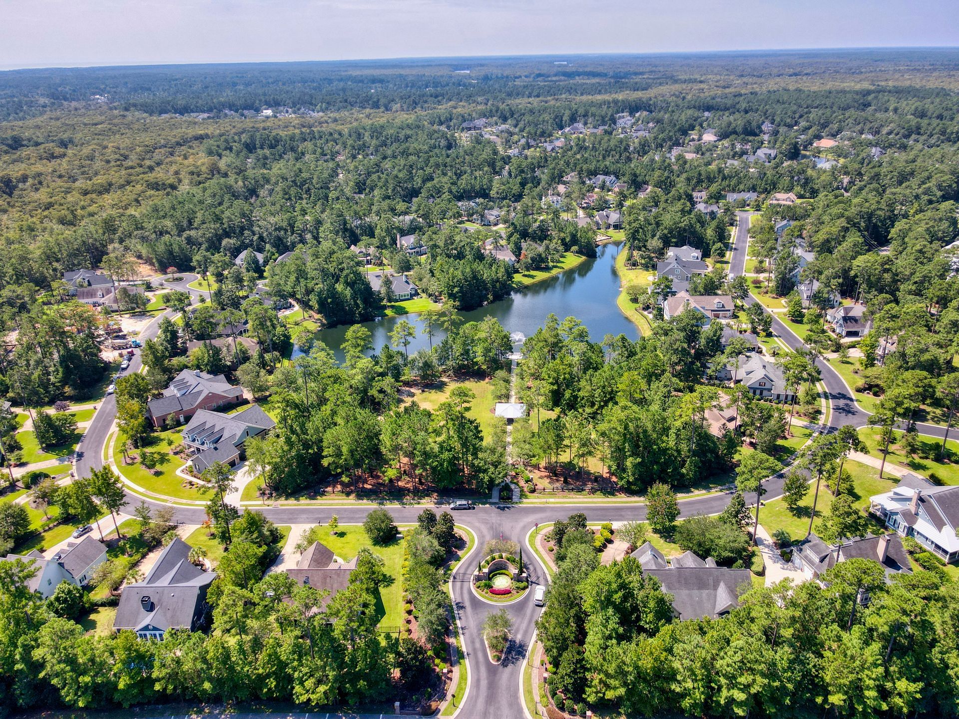 Aerial view of a suburban neighborhood with a lake, lush trees, houses, and roads on a sunny day.