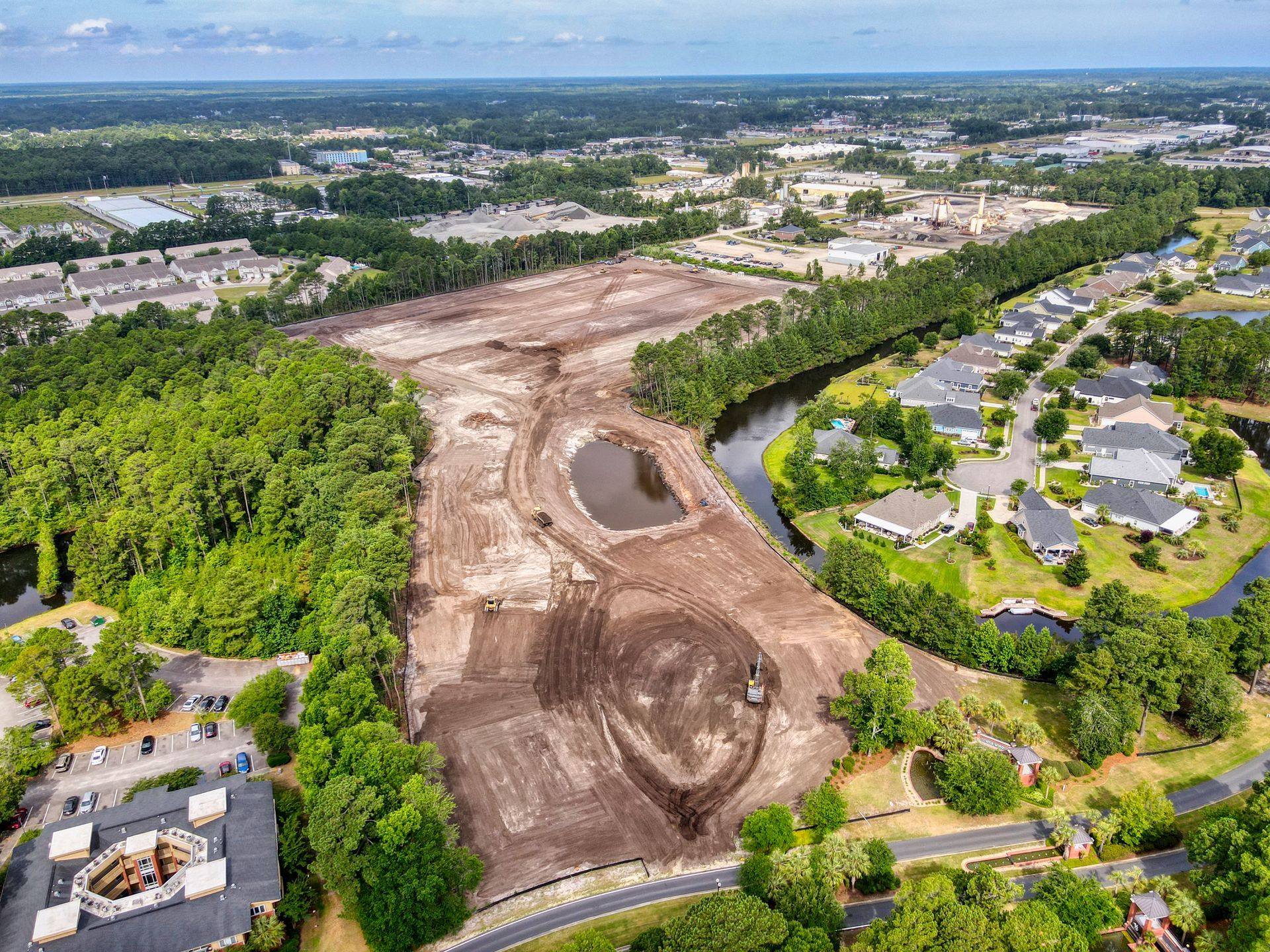 Aerial view of a cleared construction site with dirt, trees, a pond, and a residential neighborhood.