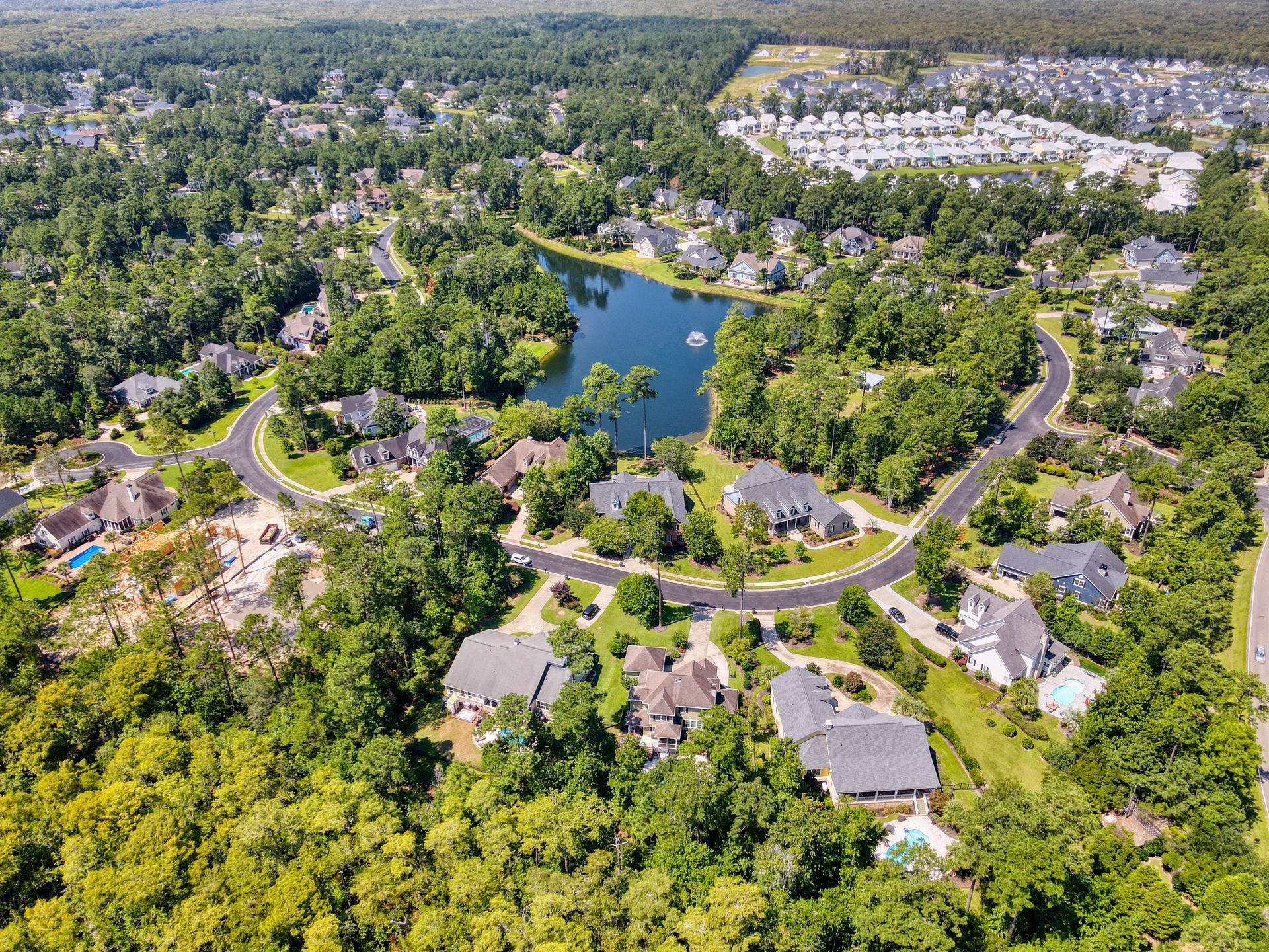 Aerial view of a suburban neighborhood with houses, a lake, trees, and curved roads under a bright sky.