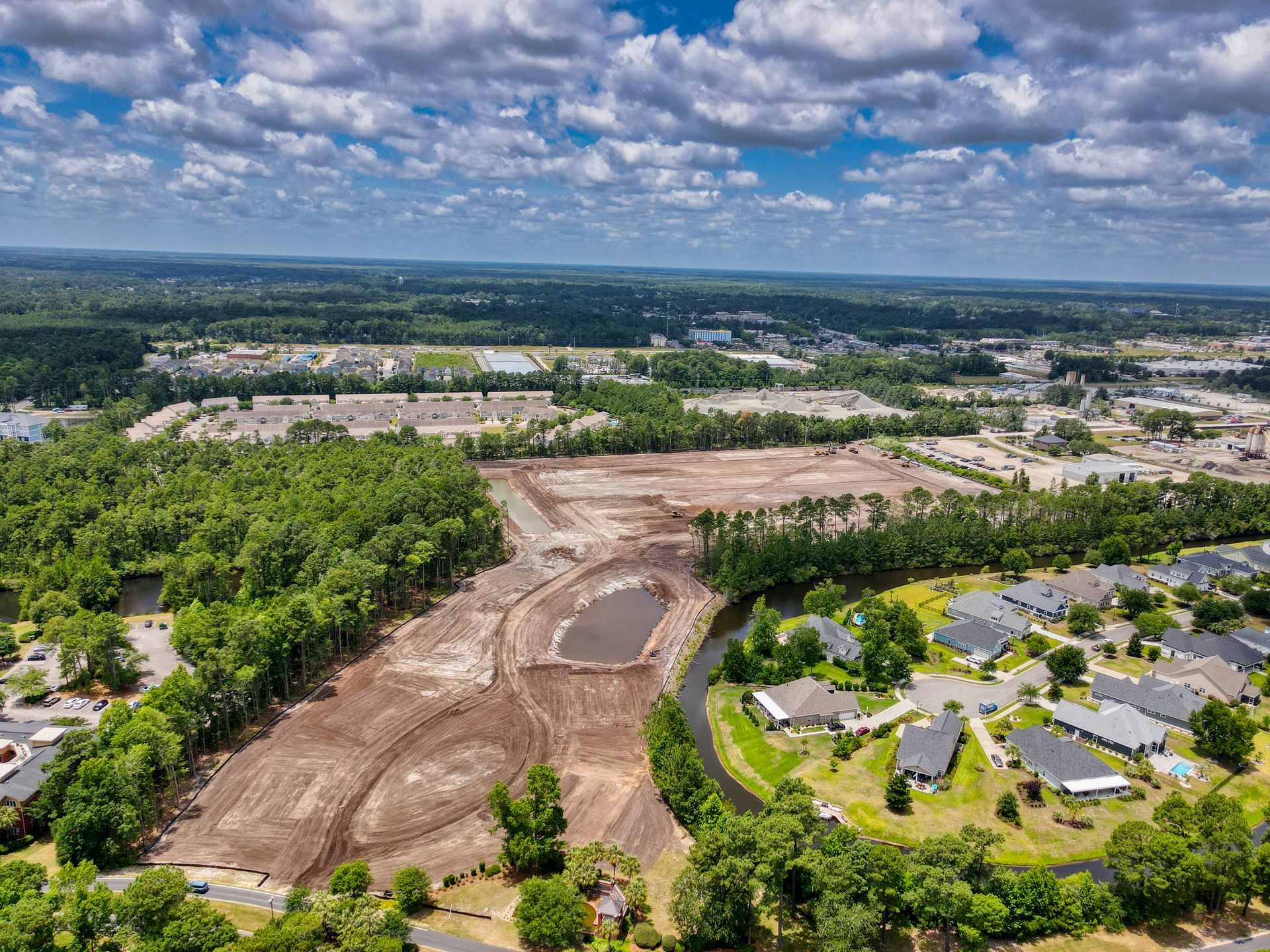 Aerial view: cleared land for development next to residential homes and a waterway; trees and clouds in the background.
