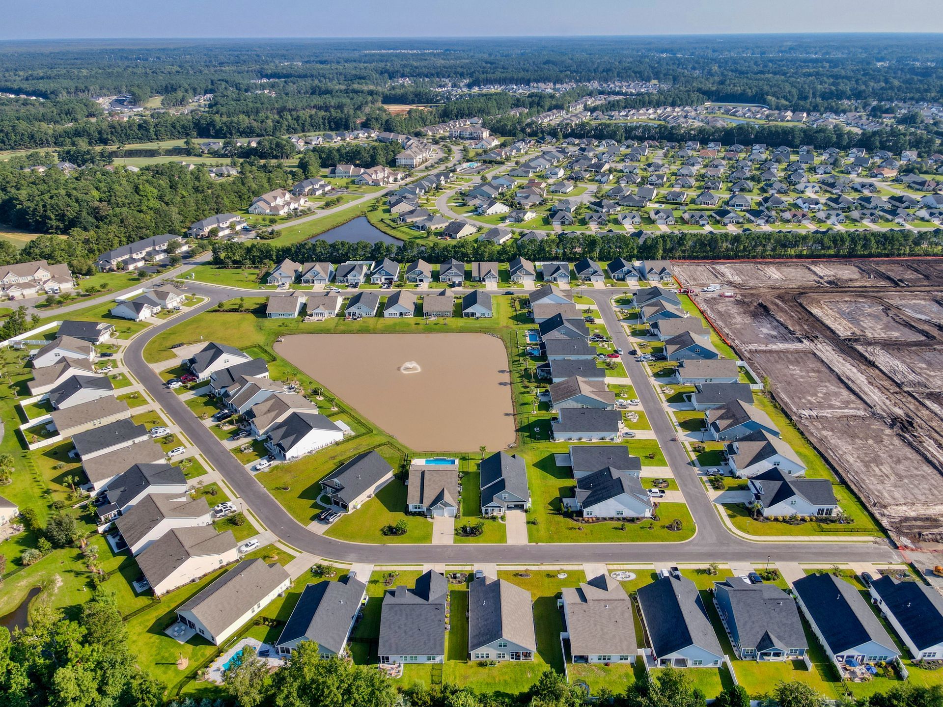 Aerial view of a suburban neighborhood with houses, streets, and a pond. Brown earth is visible to the right.