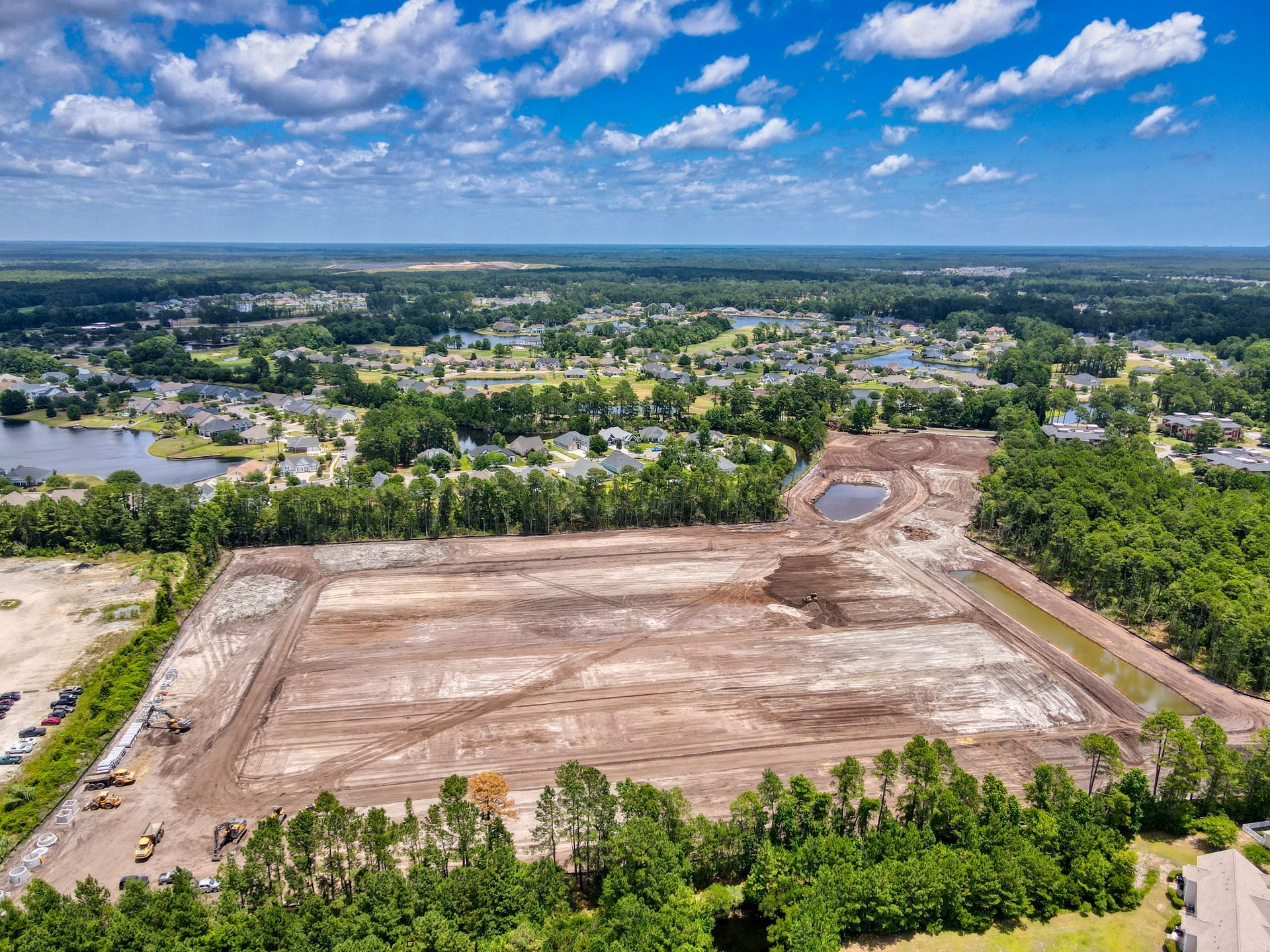 Aerial view of cleared construction site, with dirt, trees, and residential neighborhood in background.