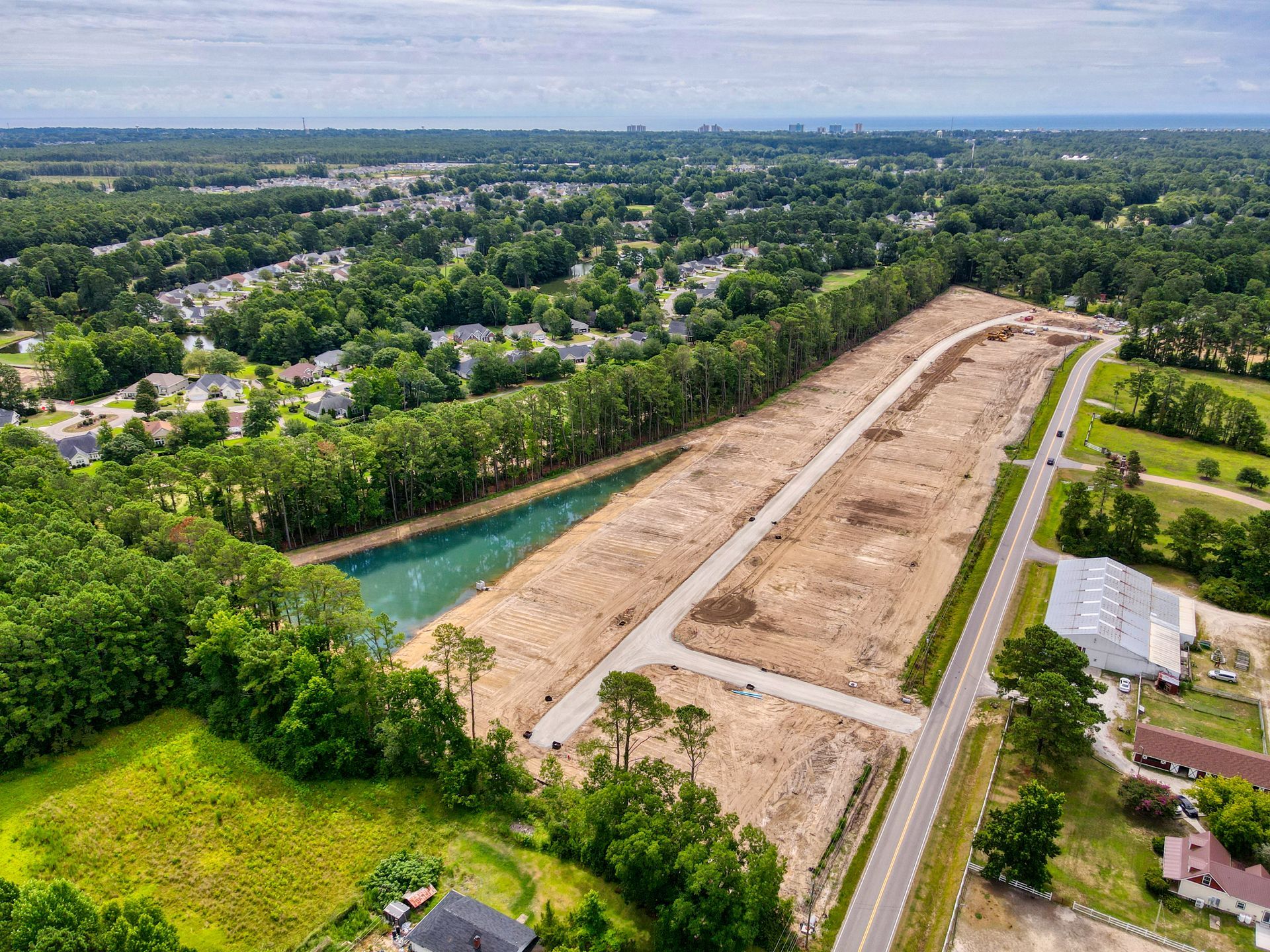 Aerial view of a cleared construction site with a long dirt road and a pond. Trees surround the area.