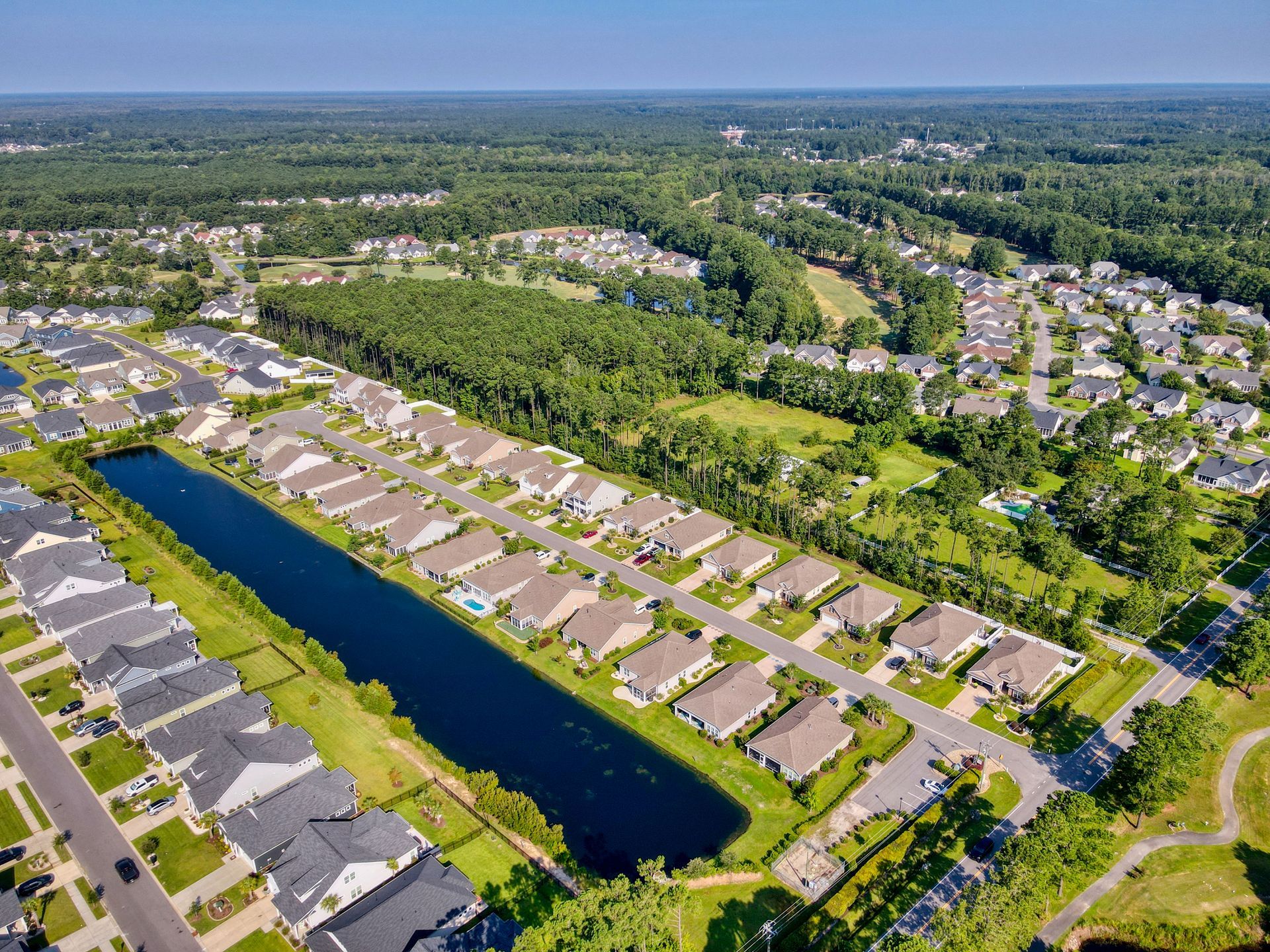 Aerial view of a suburban neighborhood with houses, a lake, green spaces, and surrounding trees under a blue sky.