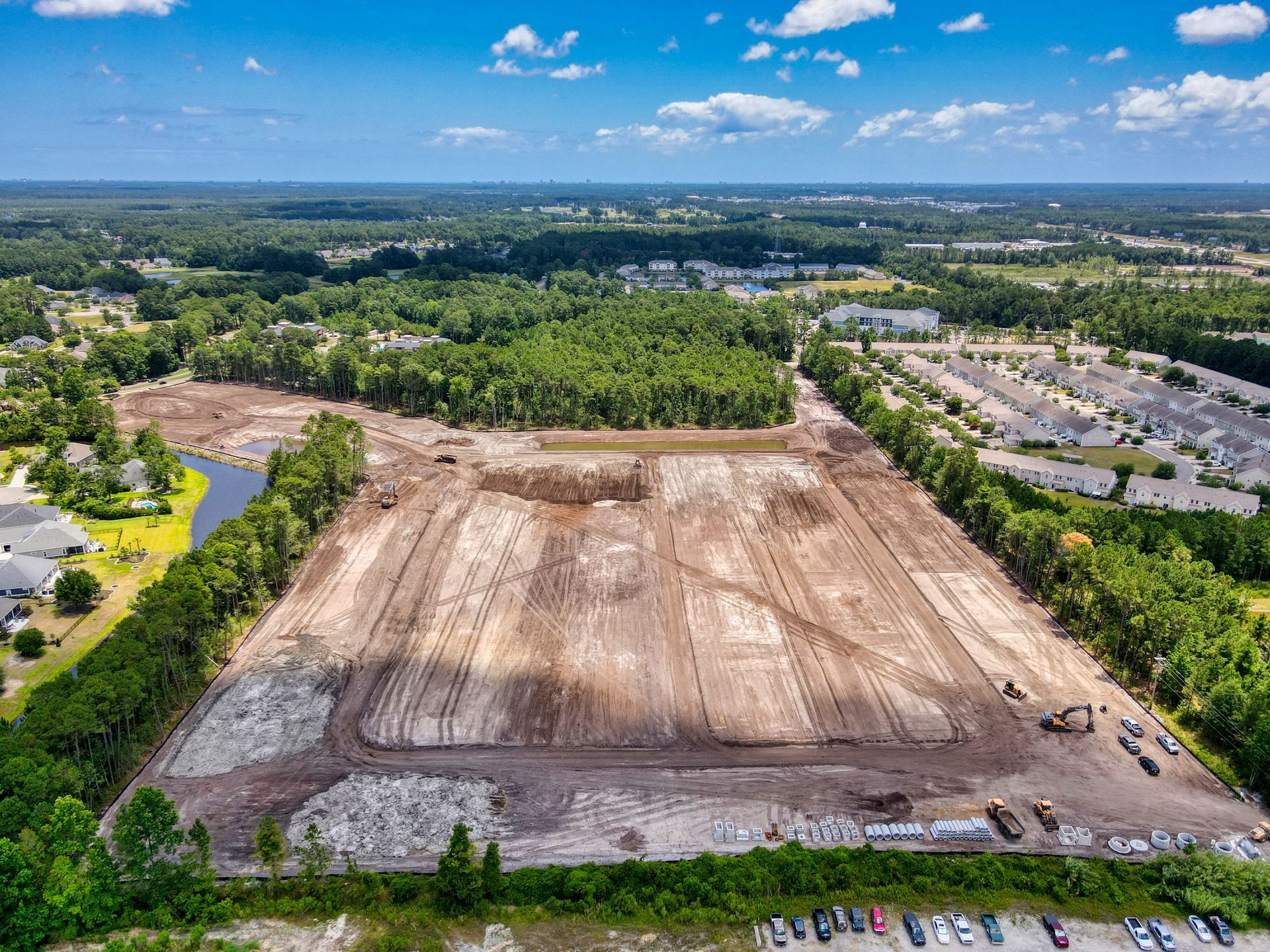 Aerial view of a cleared construction site with exposed earth, surrounded by trees and residential buildings.