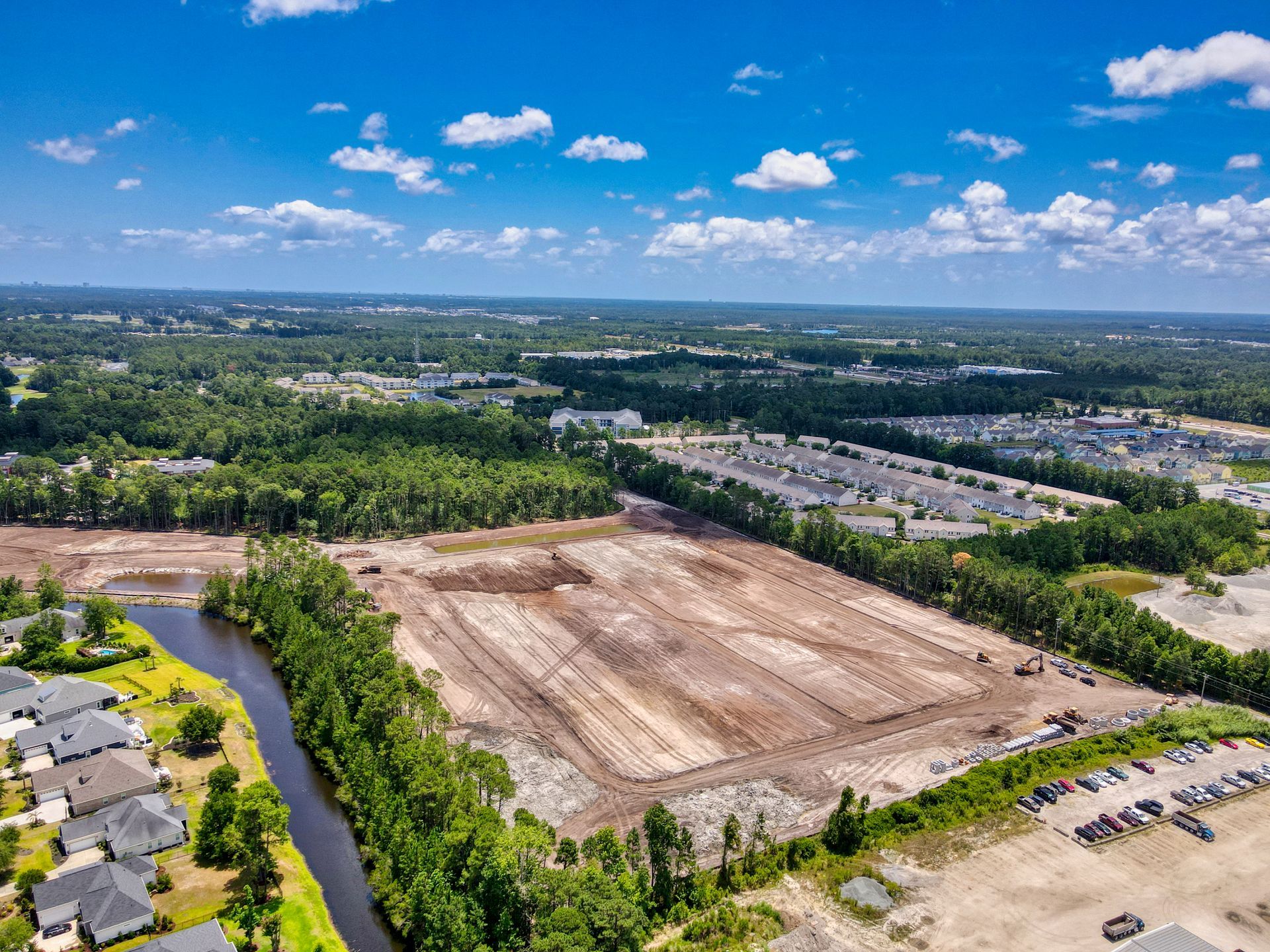 Aerial view of cleared land, bordered by trees, buildings, and a canal. Blue sky with clouds.