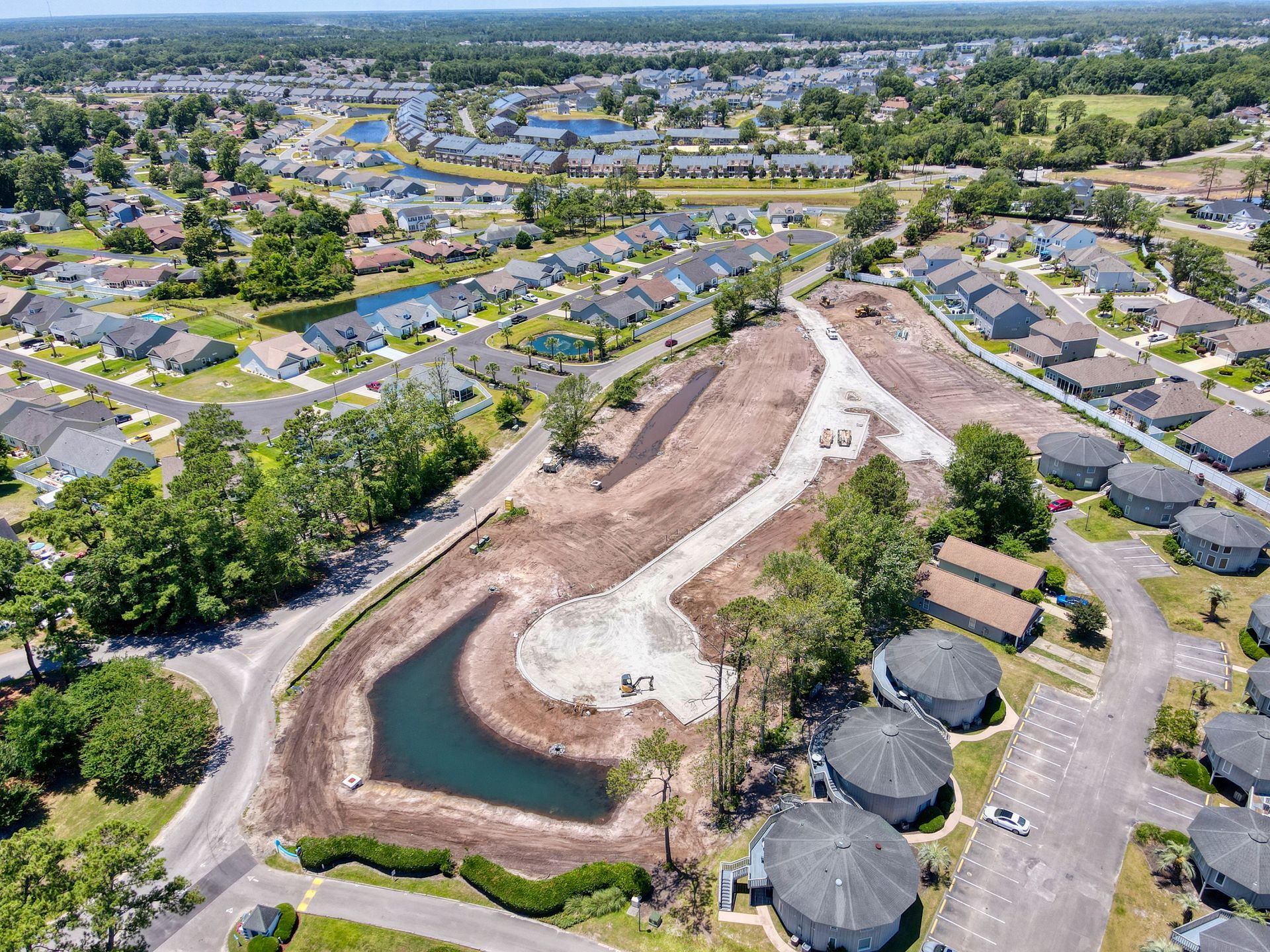 Aerial view of a construction site with a pond, surrounded by residential houses and green trees.