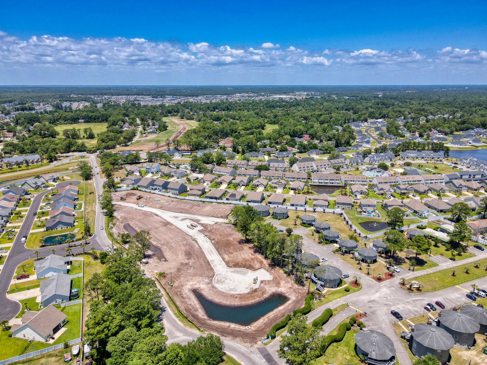 Aerial view of a suburban neighborhood with a construction site and retention pond. Green trees, blue sky.