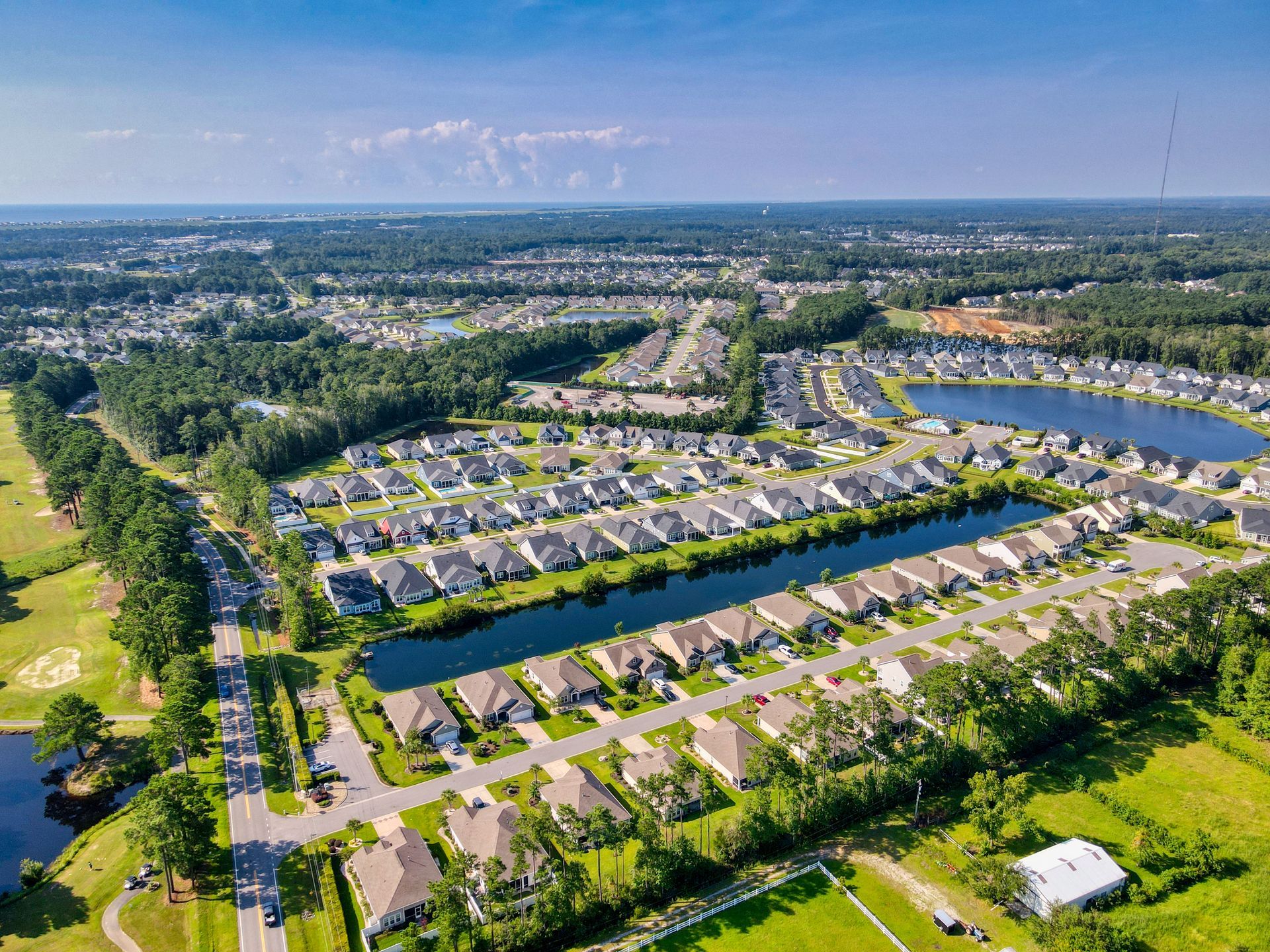 Aerial view of a suburban neighborhood with houses, a lake, green spaces, and trees under a clear blue sky.