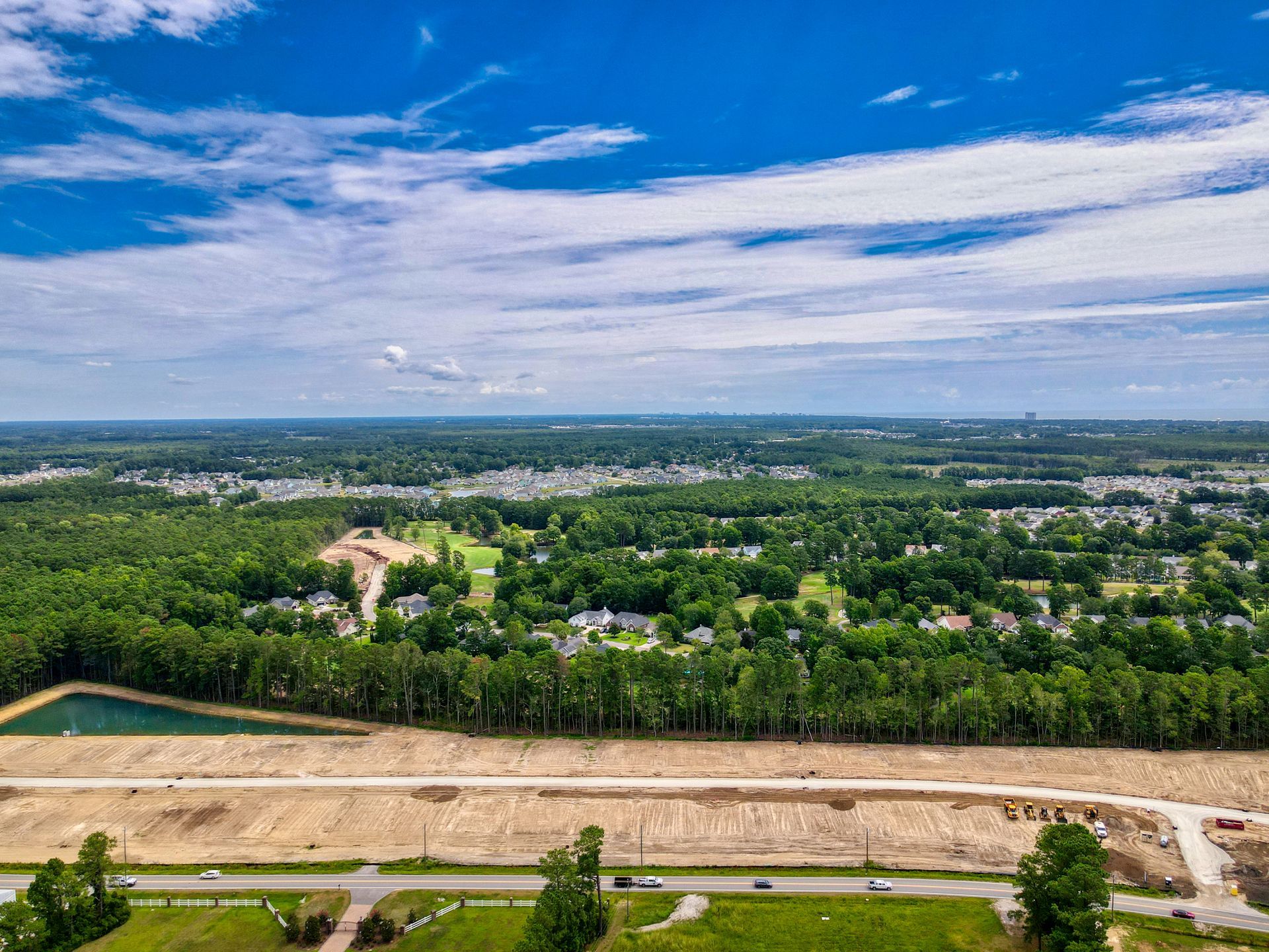 Aerial view of land clearing, green trees, houses, and cloudy blue sky.