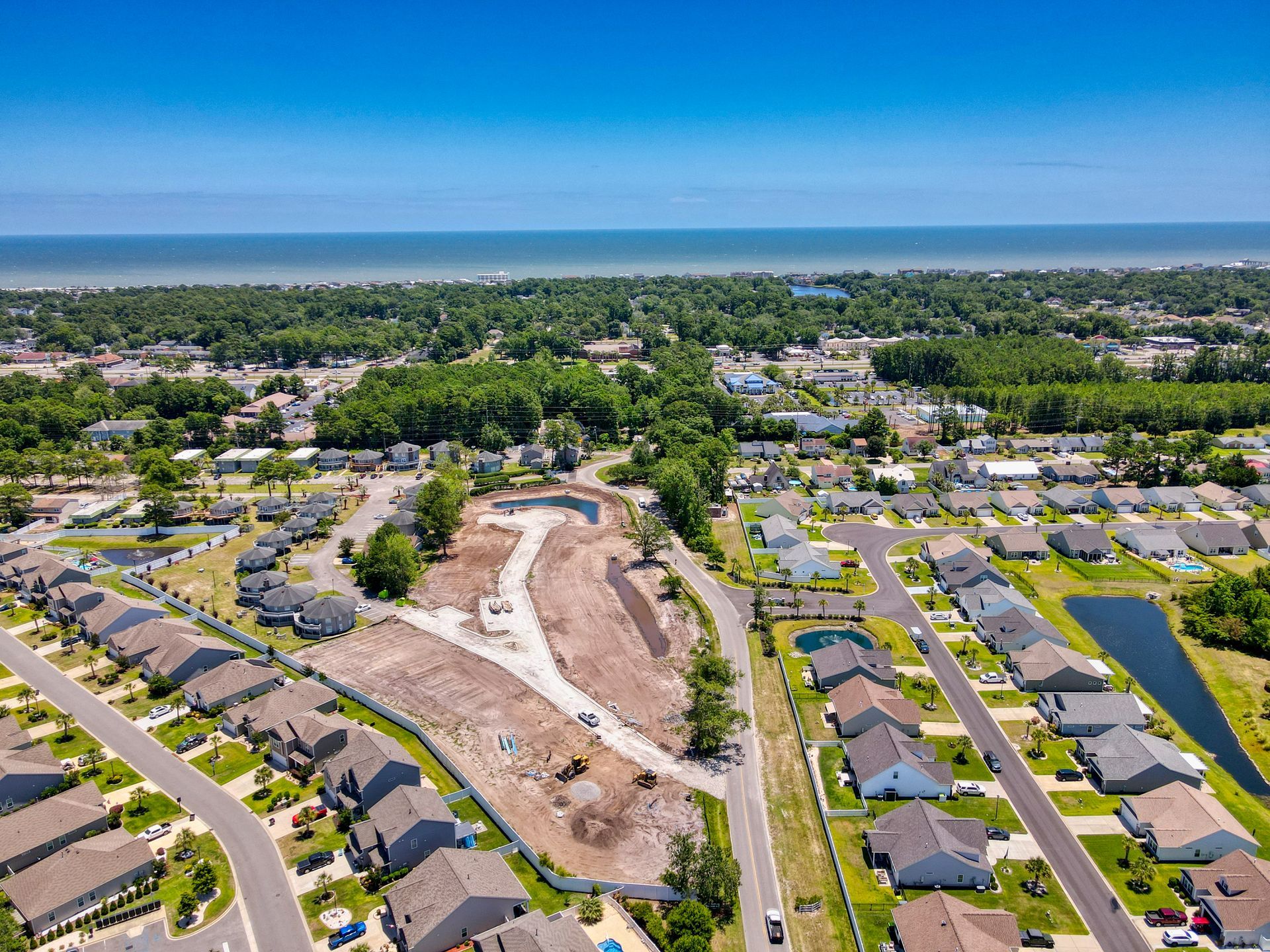Aerial view of a coastal town with houses, construction site, trees, and the ocean in the distance under a blue sky.