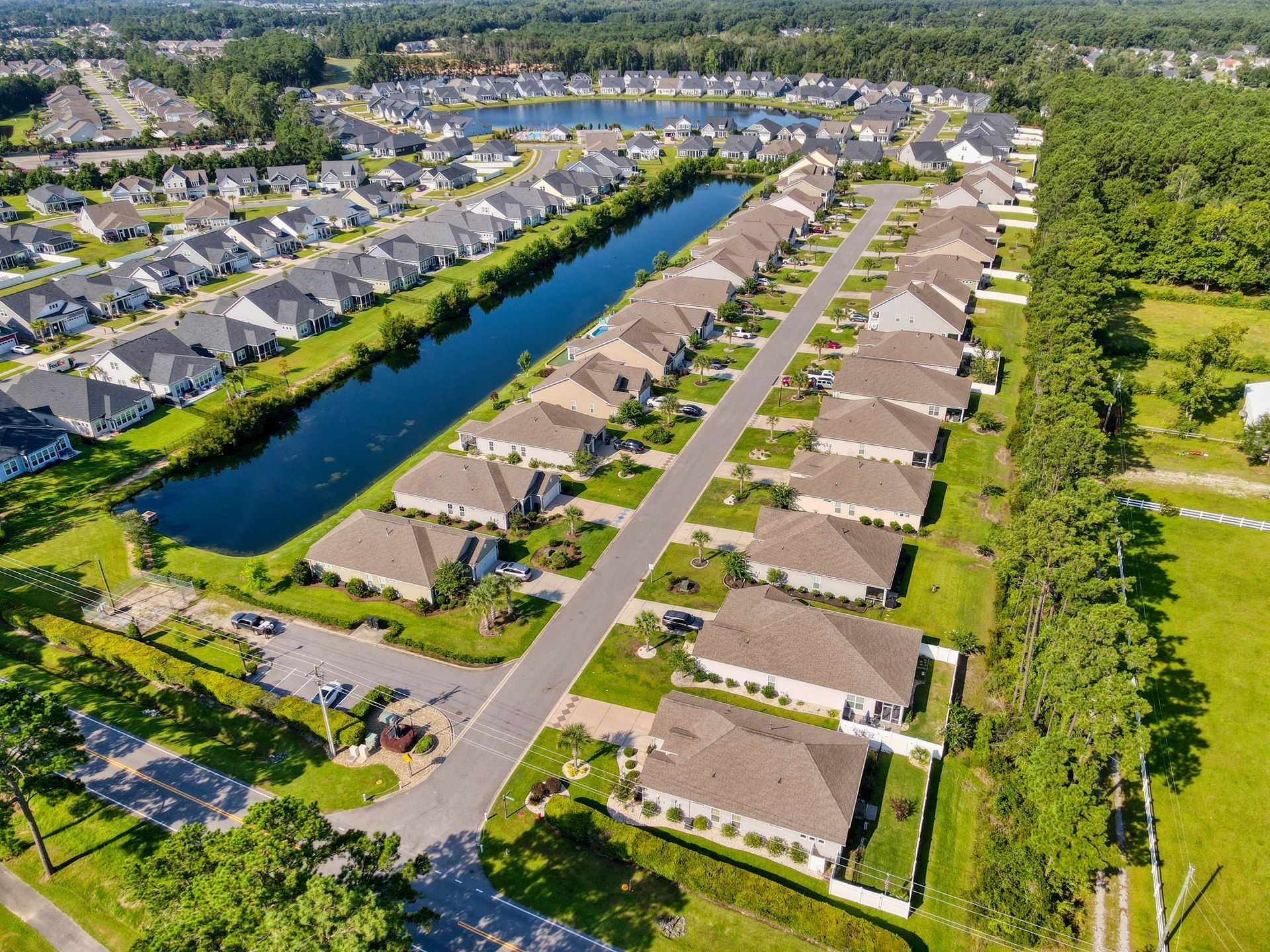 Aerial view of a residential neighborhood with houses lining a street and a lake. Green grass and trees surround the houses.