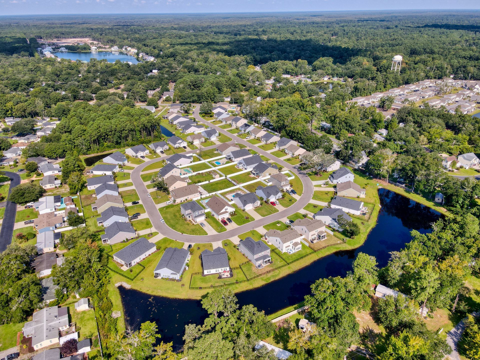 Aerial view of a suburban neighborhood with houses surrounding a pond and a curved road, surrounded by trees.