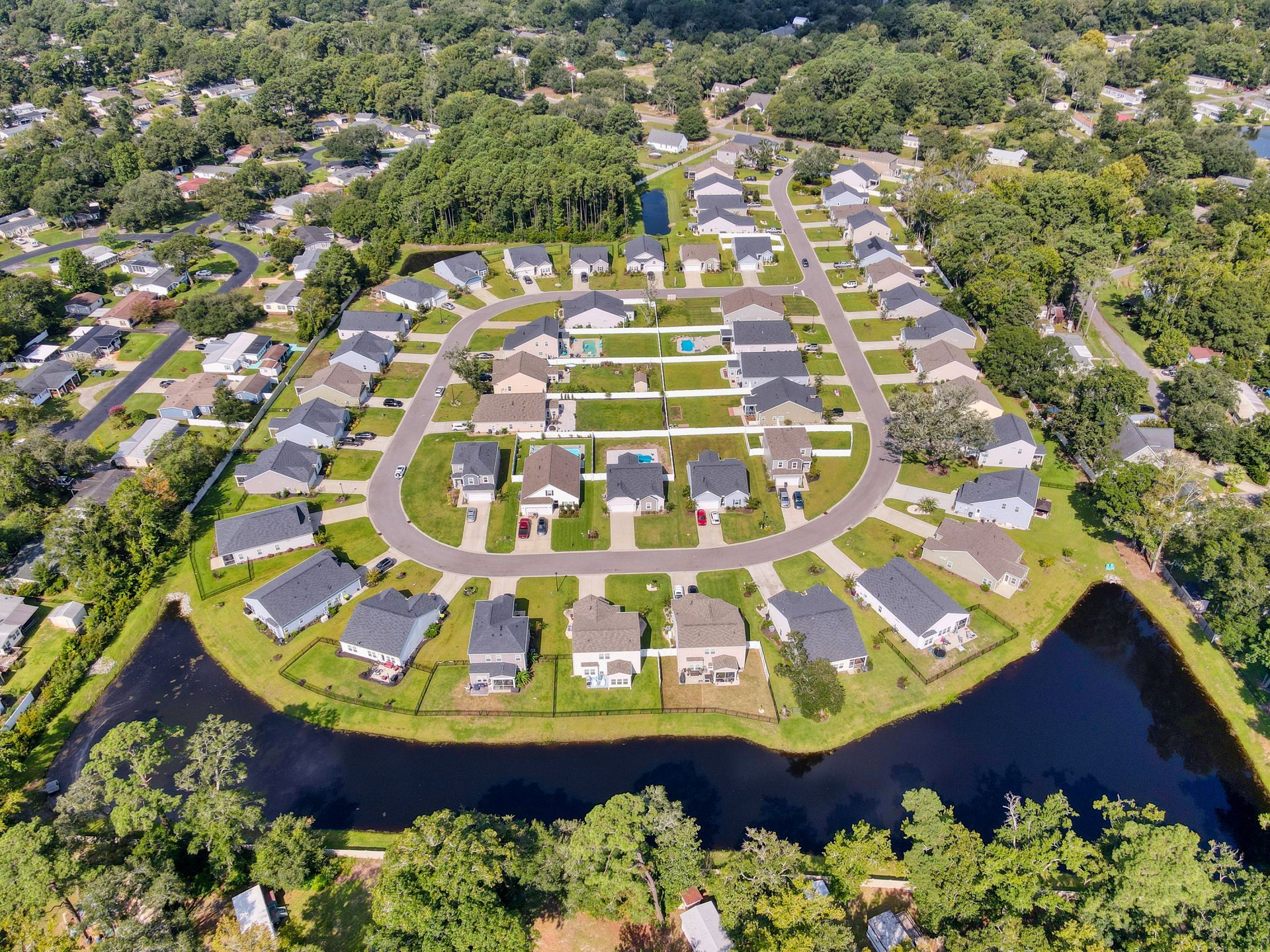 Aerial view of a residential neighborhood with houses arranged around a curved street, bordered by a pond and greenery.