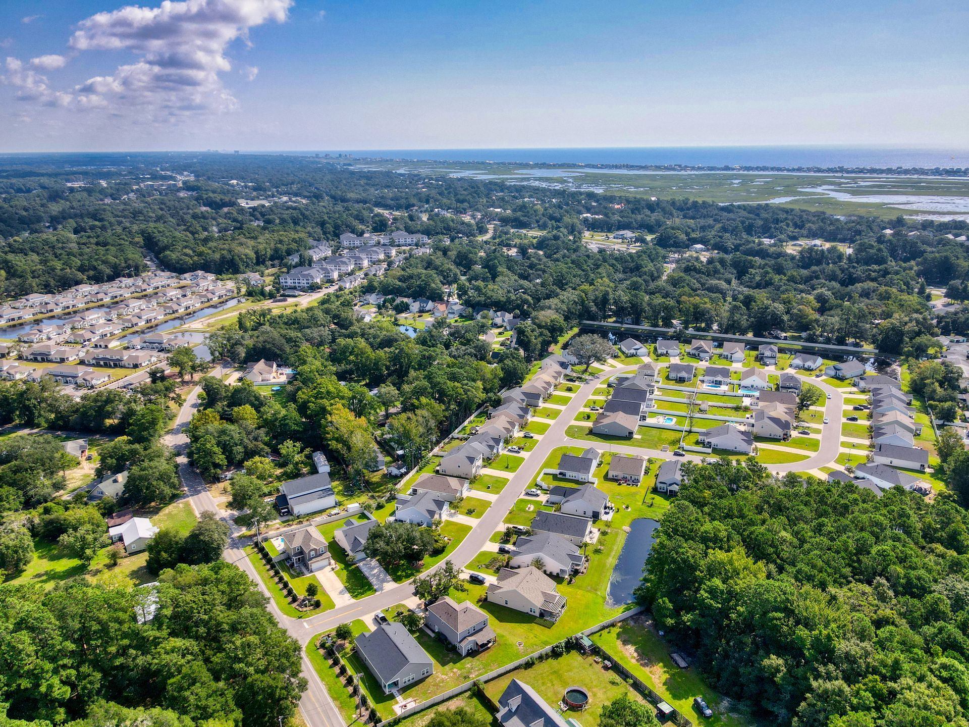 Aerial view of suburban neighborhood with houses, trees, roads, and a body of water in the background under a blue sky.