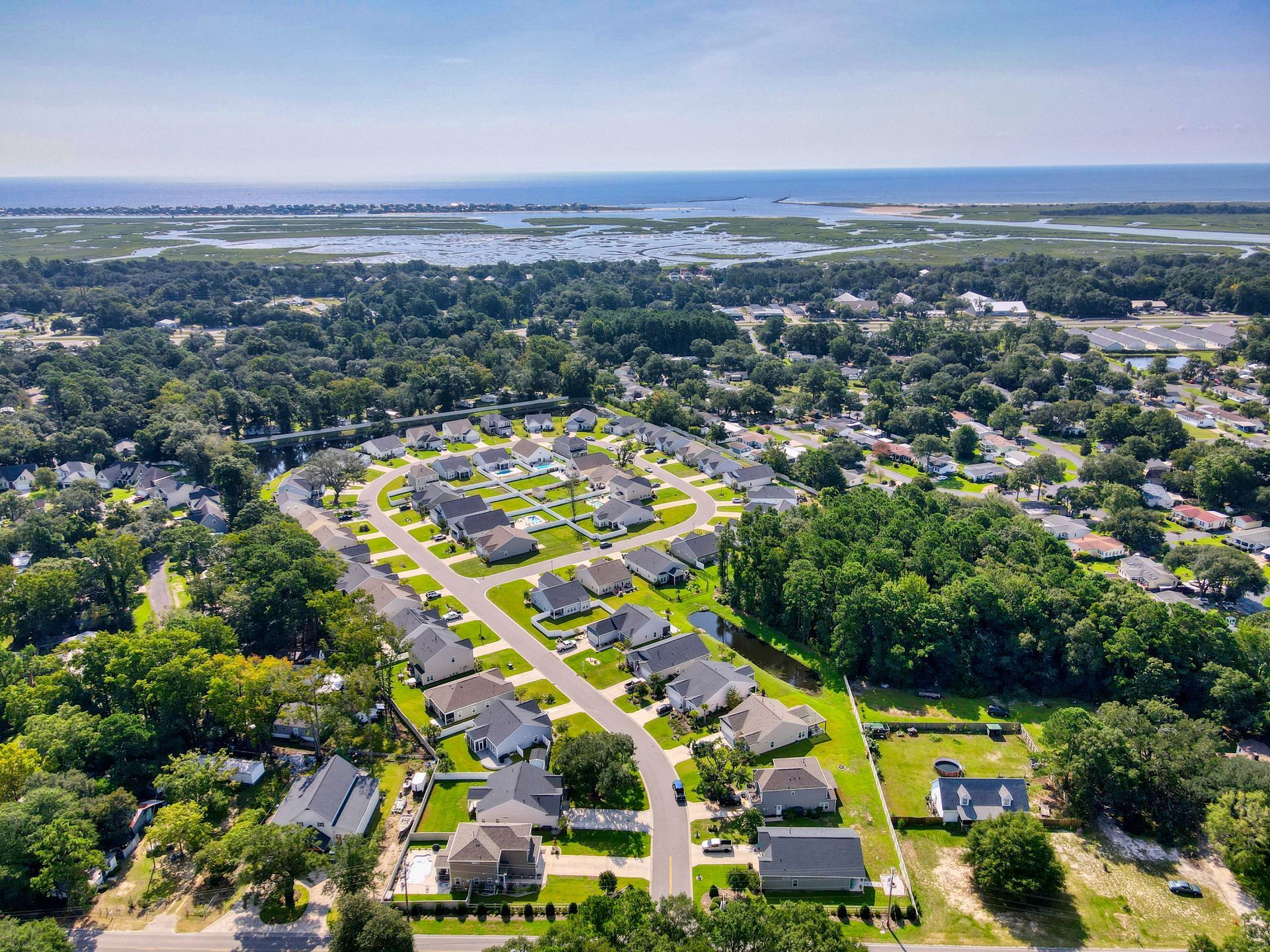 Aerial view of a suburban neighborhood with houses, roads, trees, and water in the background on a sunny day.