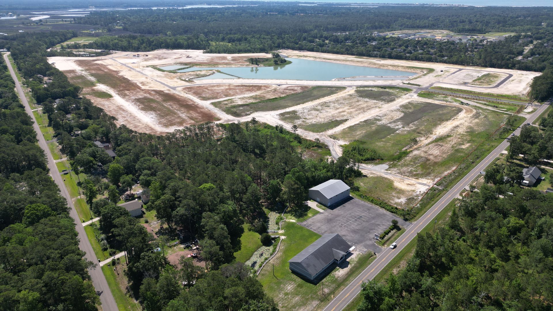 Aerial view of a cleared construction site with graded land, a retention pond, and surrounding trees.
