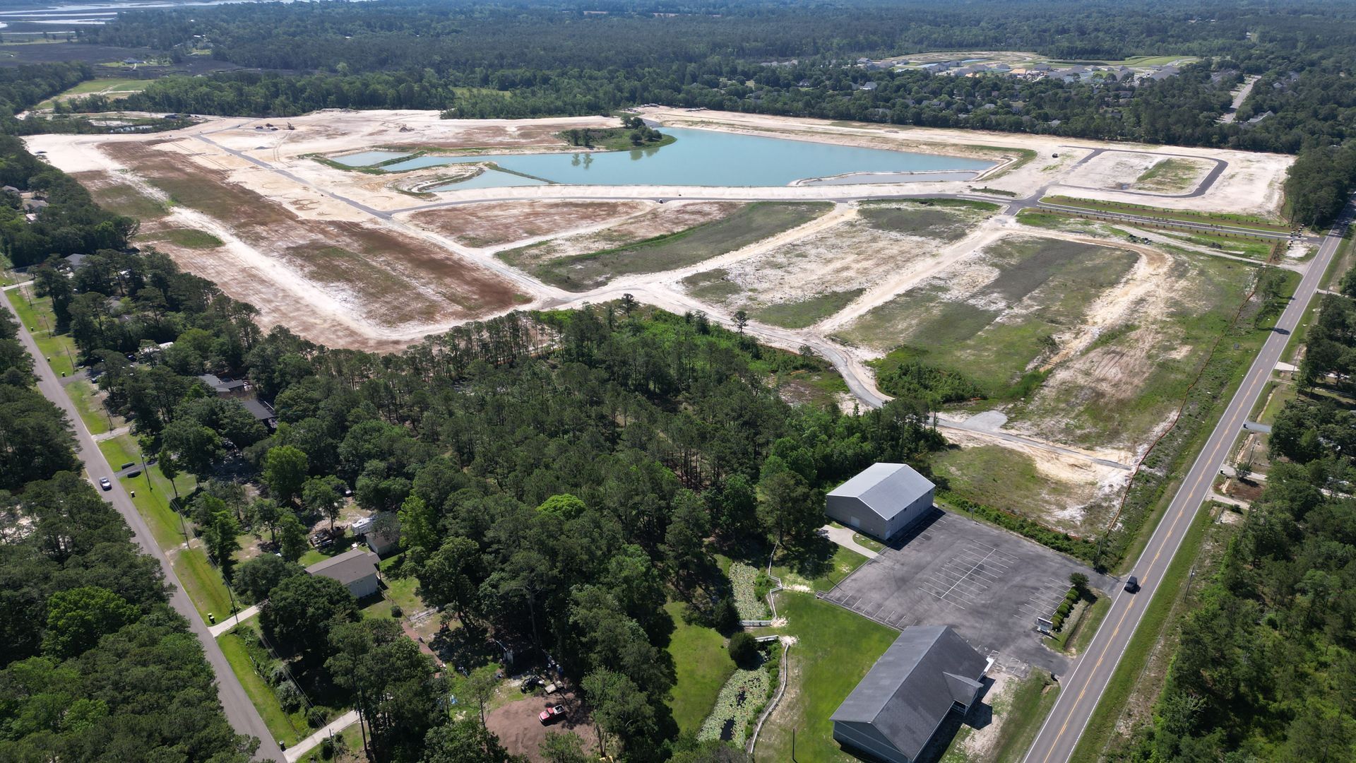 Aerial view of a cleared construction site with a pond and access road, surrounded by trees.