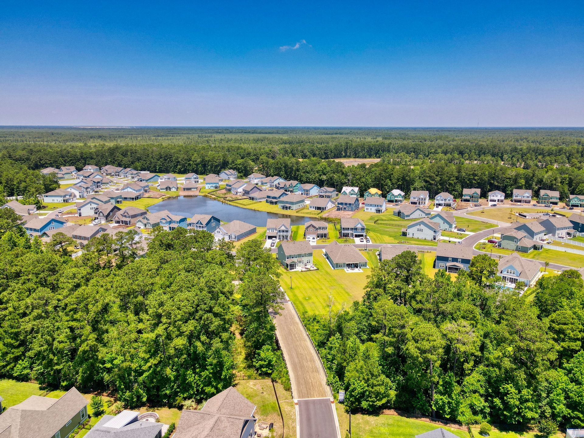 Aerial view of a suburban neighborhood with houses, a lake, and trees under a clear blue sky.