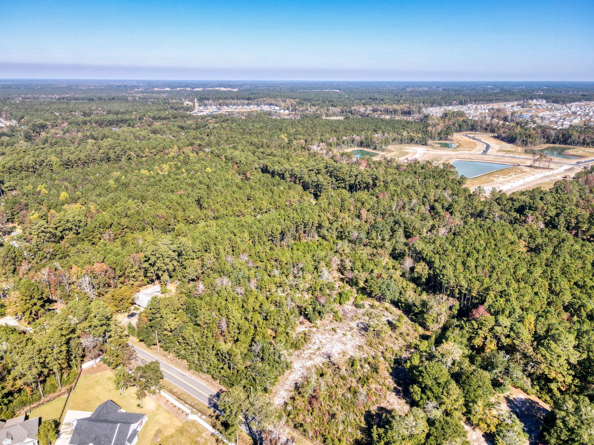 Aerial view of a forested area with a road running through it and a glimpse of houses and water features in the distance.
