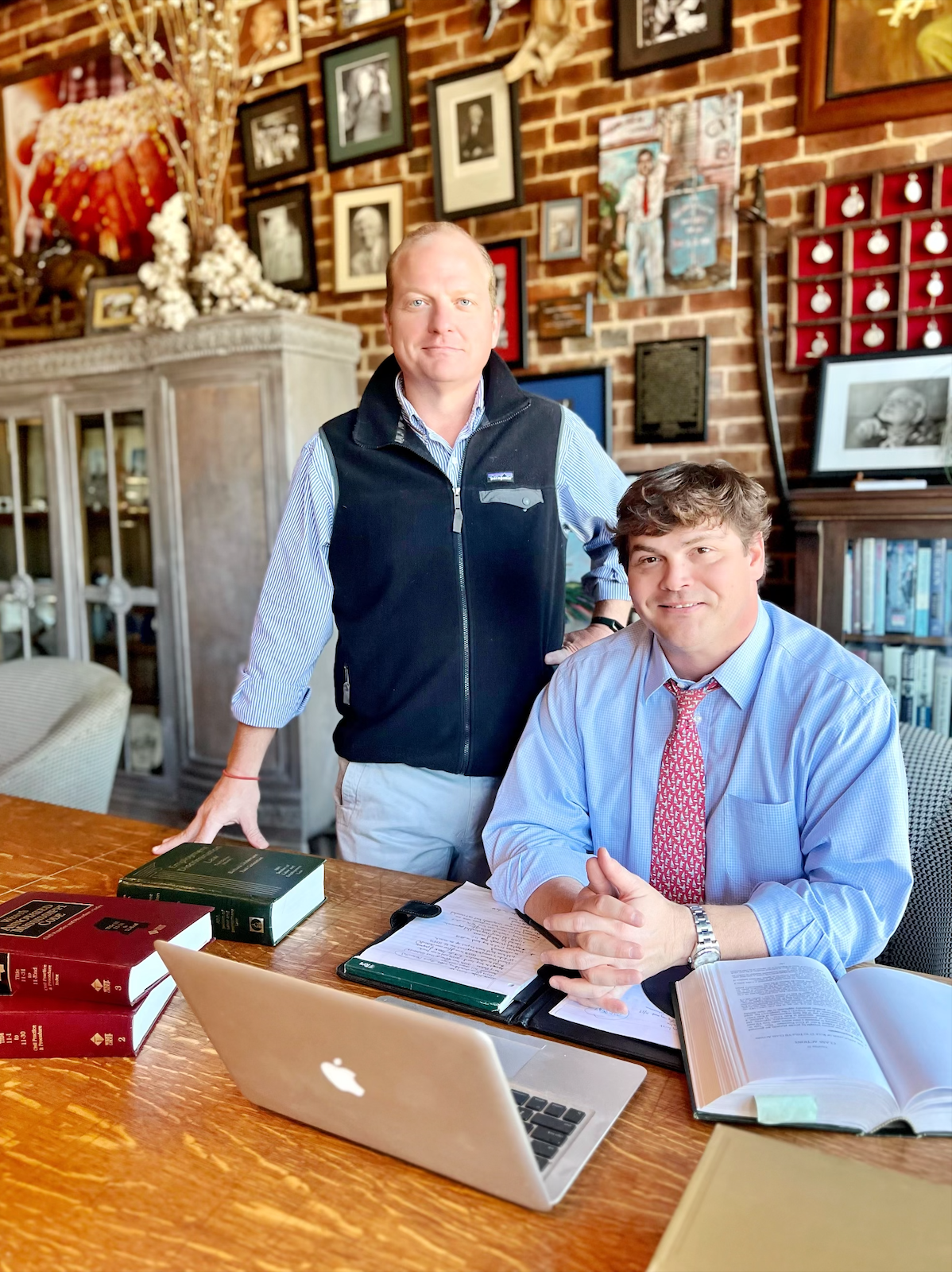 Two men at a wooden desk with books, a laptop, and papers, in a room with brick wall and photographs.