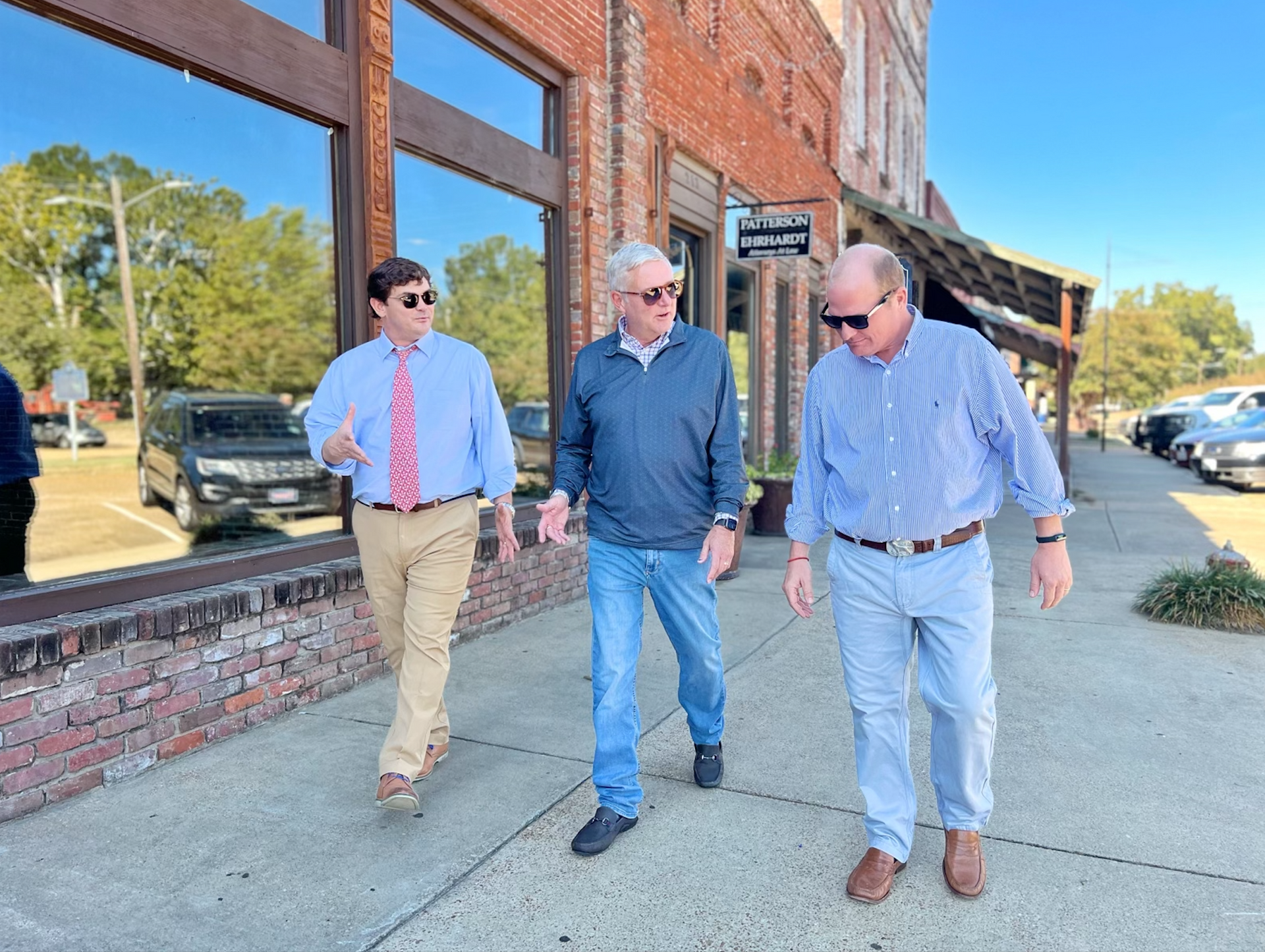 Three men walking and talking on a sidewalk in front of a brick building.