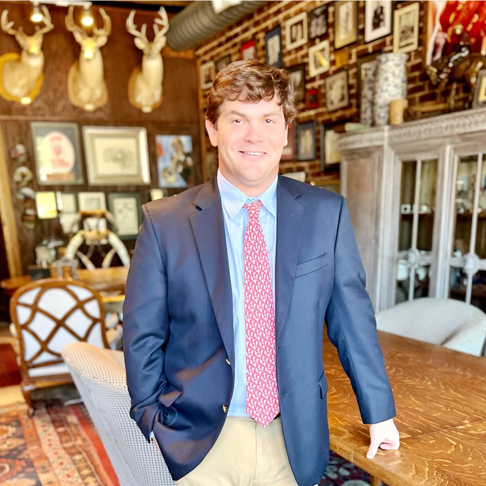 Man in a navy blazer and patterned tie stands near a wooden table; many framed pictures hang on the wall.