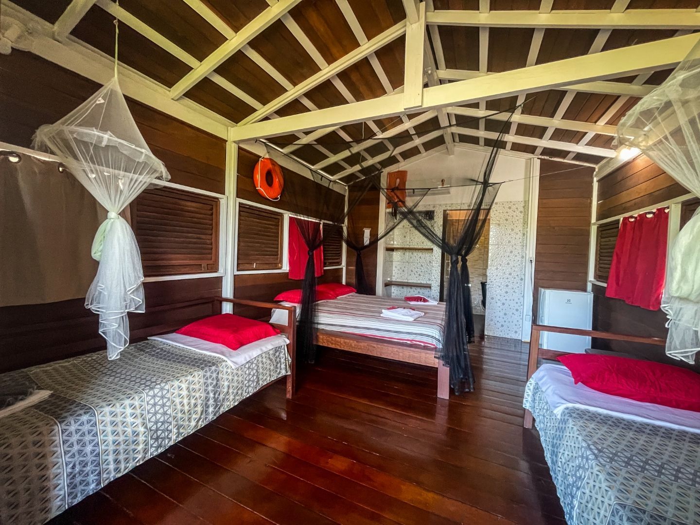 Interior de cabana de madeira com três camas, mosquiteiros, detalhes em vermelho e piso de madeira.