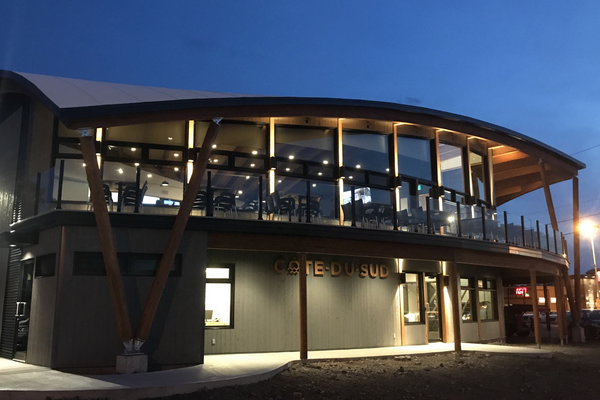 Un restaurant moderne à deux étages avec des fenêtres en verre incurvées et un surplomb en bois au crépuscule.
